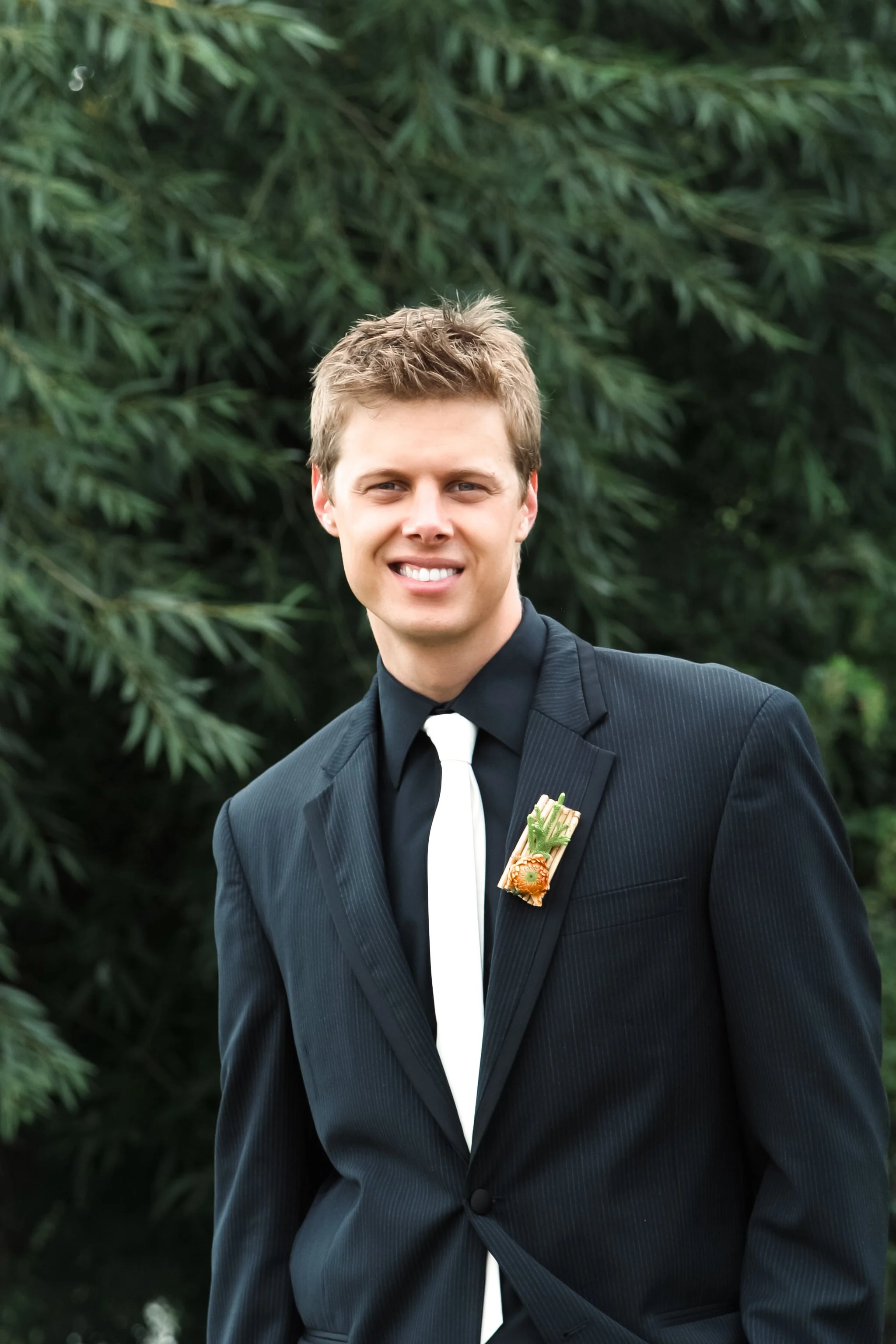 A young man with light brown hair wearing a black suit, white shirt, and black tie, standing outdoors with greenery in the background. He has a floral boutonniere on his lapel and is smiling.
