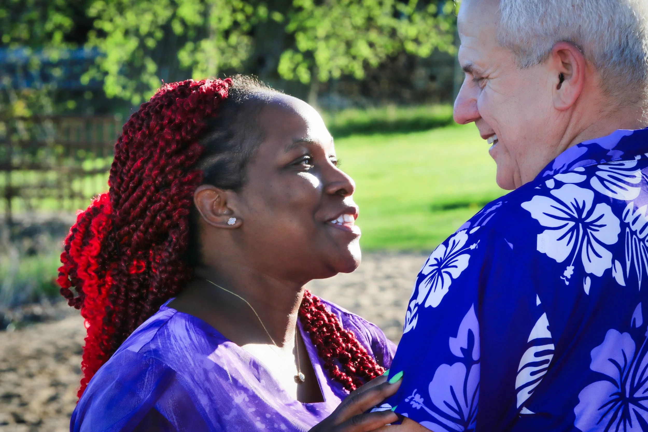 A woman with braided red hair and a man with gray hair smiling at each other outdoors in a park with green trees and grass.