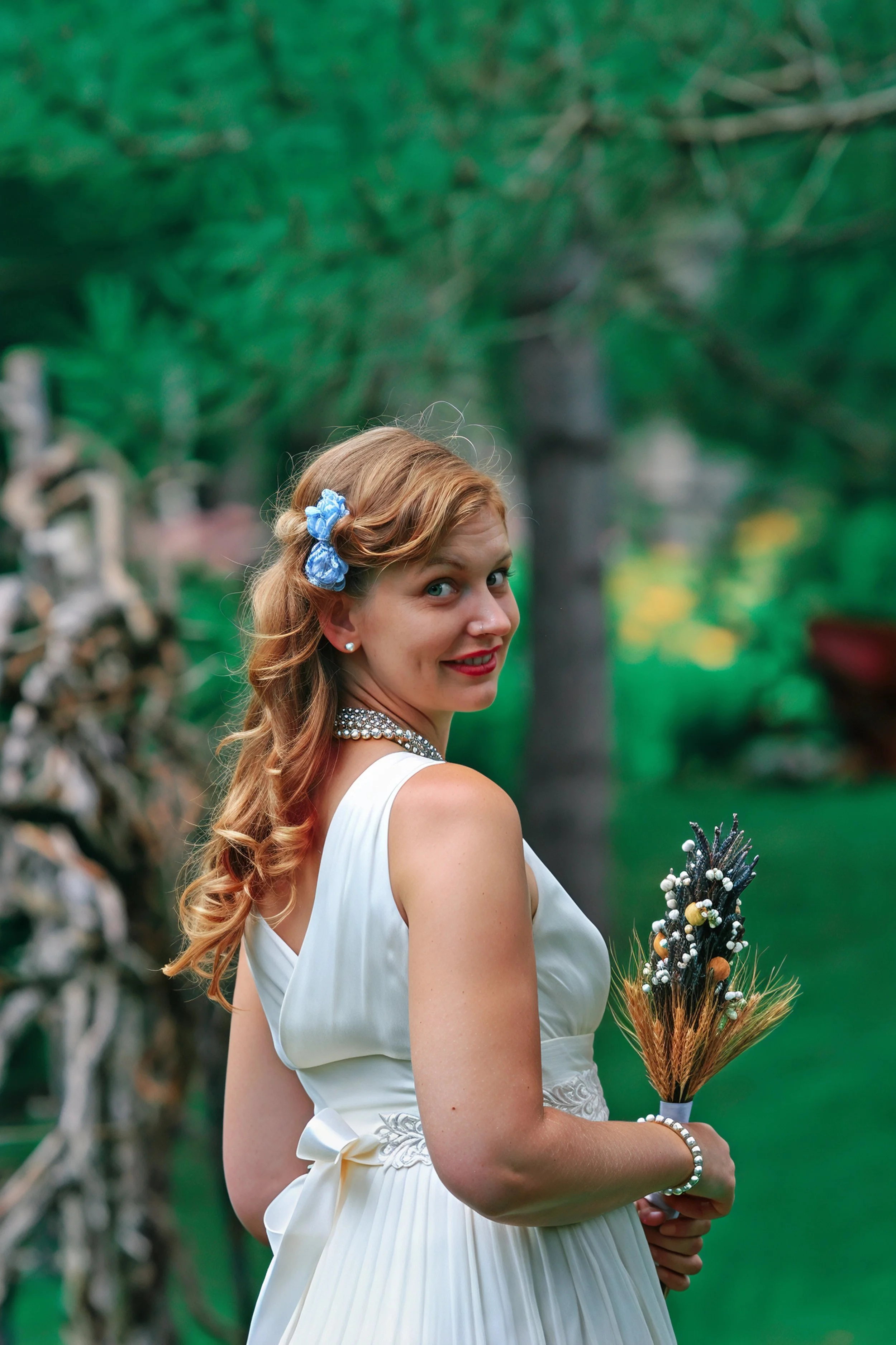 A young woman in a white dress holding a bouquet, with a blue flower in her hair, standing outdoors with a green forest background.
