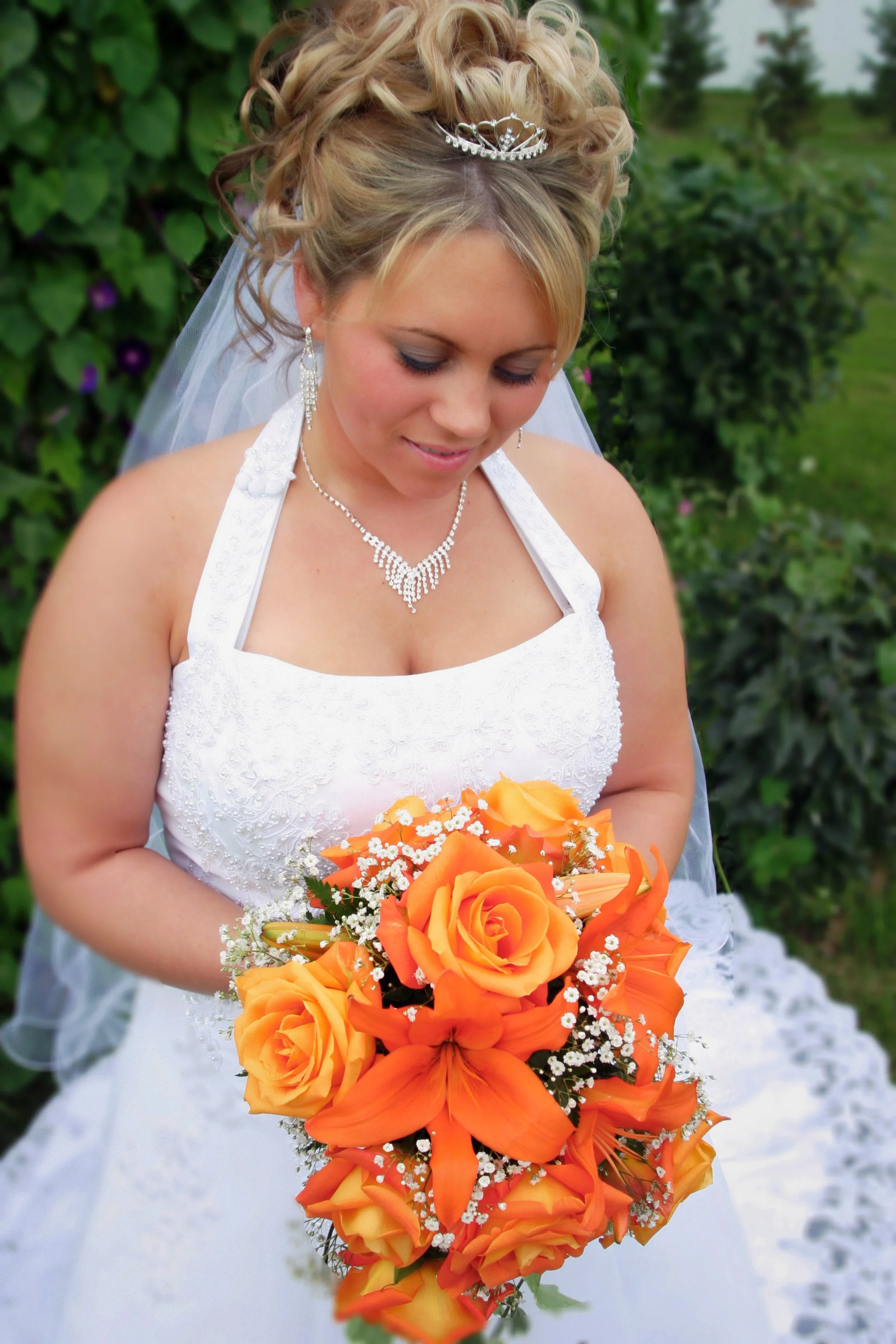 Close-up of a bride holding a bouquet of orange roses, lilies, and white baby's breath, wearing a wedding dress, jewelry, and a tiara, with greenery in the background.