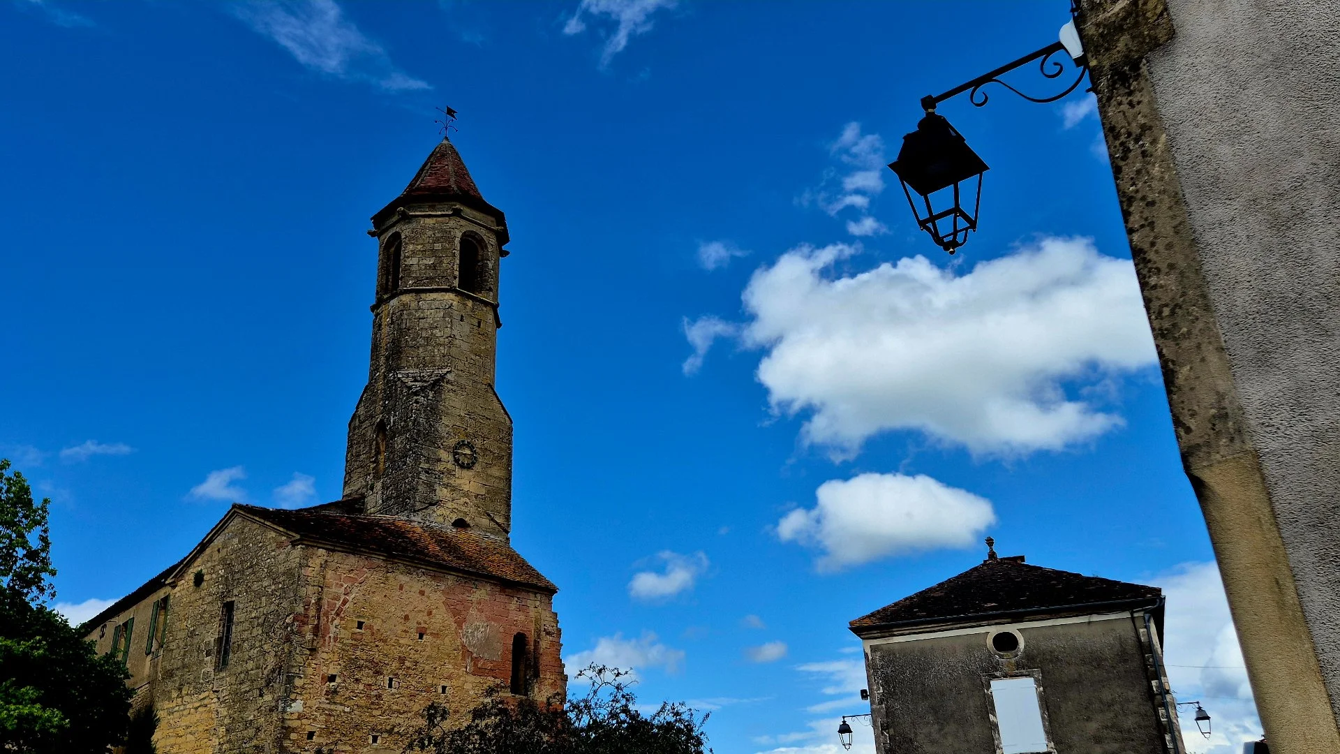 A picture of one of the Belves Bell Towers. Old stone church with a tall bell tower and a weather vane on top, black lantern attached to a building on the right, clear blue sky with some white clouds.