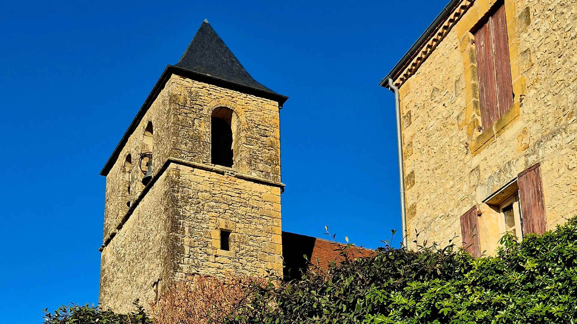 A picture of a belltower in Marnac Dordogne with a beautiful old stone house next door. The sky is bright blue and clear.