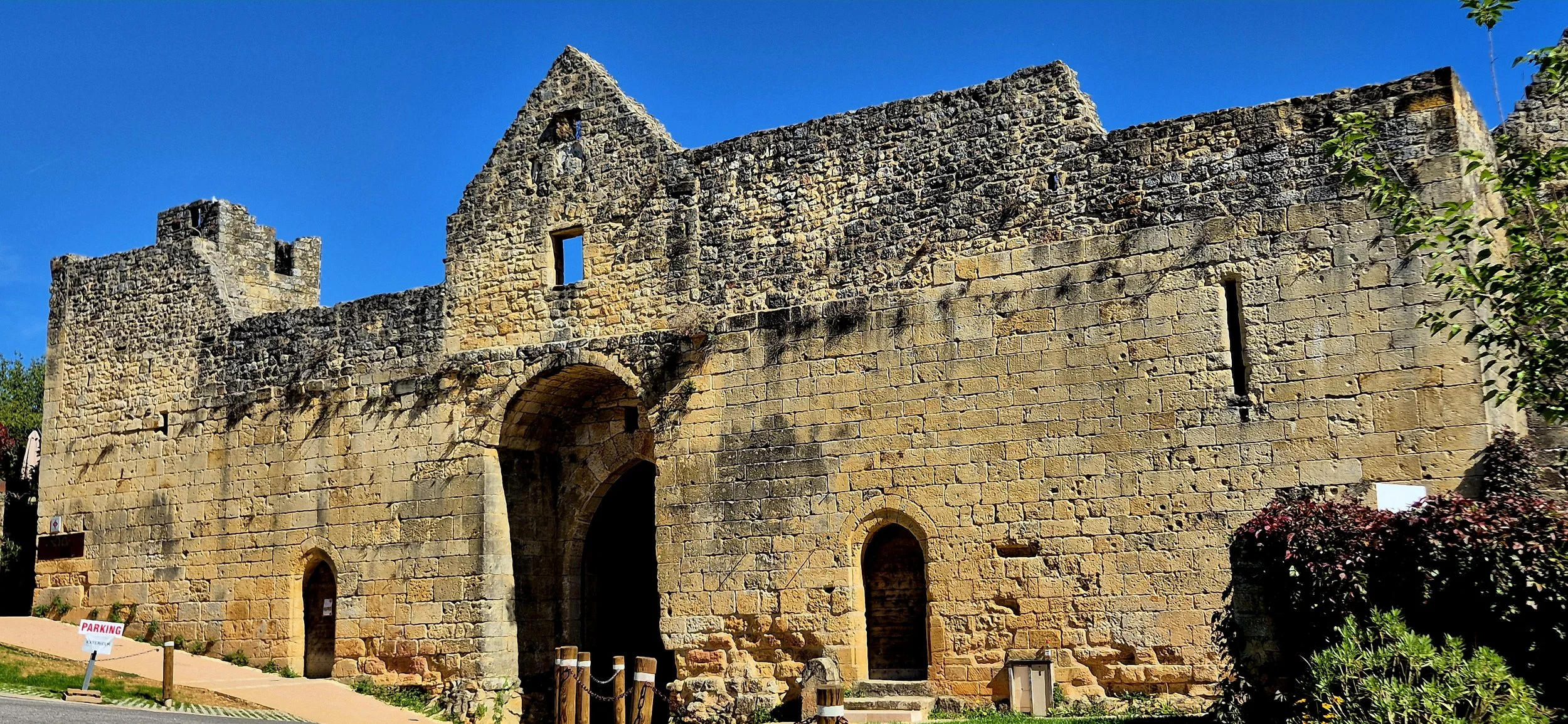 The image shows an old stone structure, possibly a castle or fort, with a large archway and small windows. The sky is clear and blue, and there are some bushes with pink flowers and a small sign indicating parking in front of the structure.