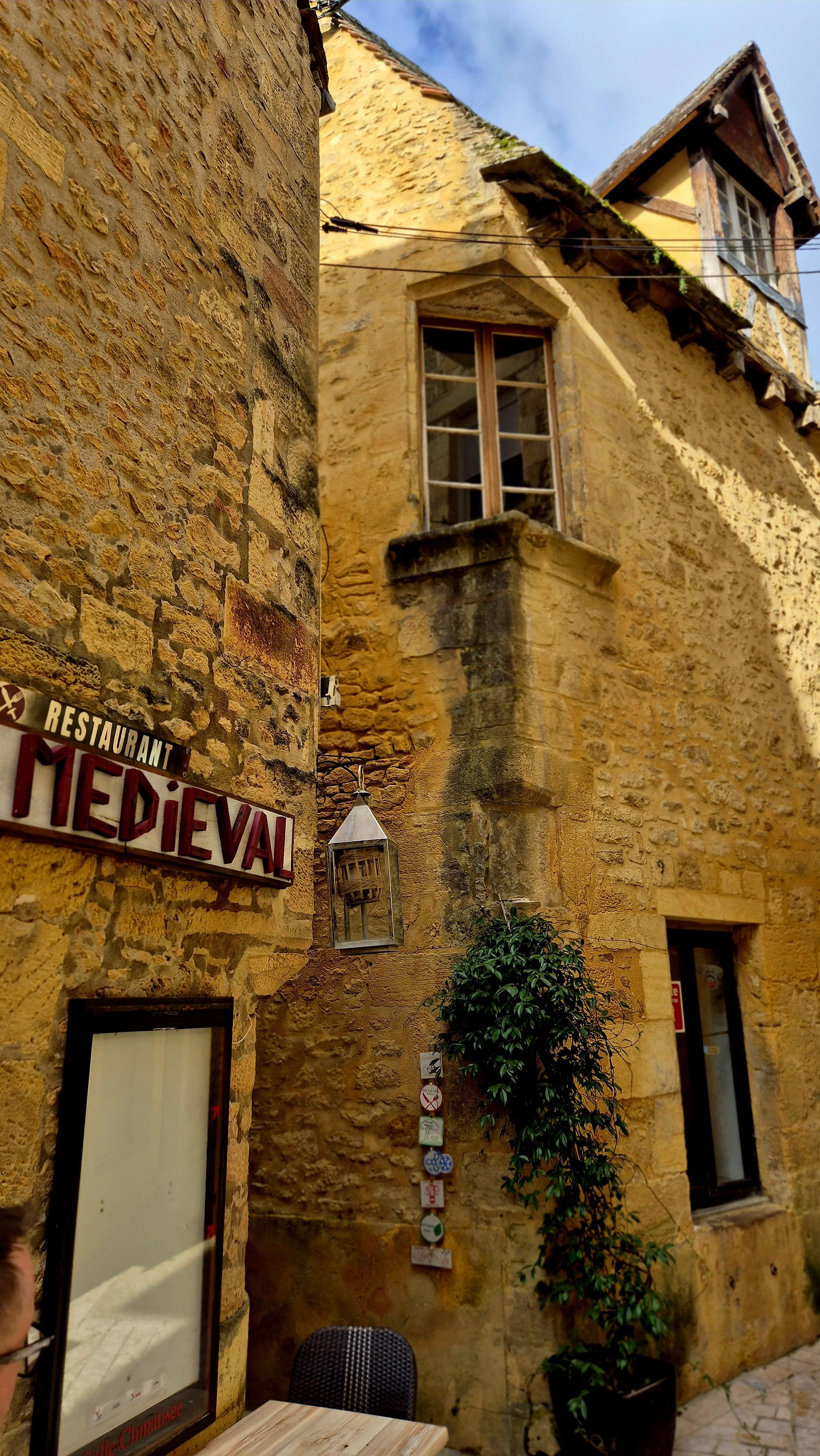 A yellow stone building with a window, a sign that reads "HIEDIEVAL," a lantern, potted plant, and outdoor seating area beneath a partly cloudy sky.