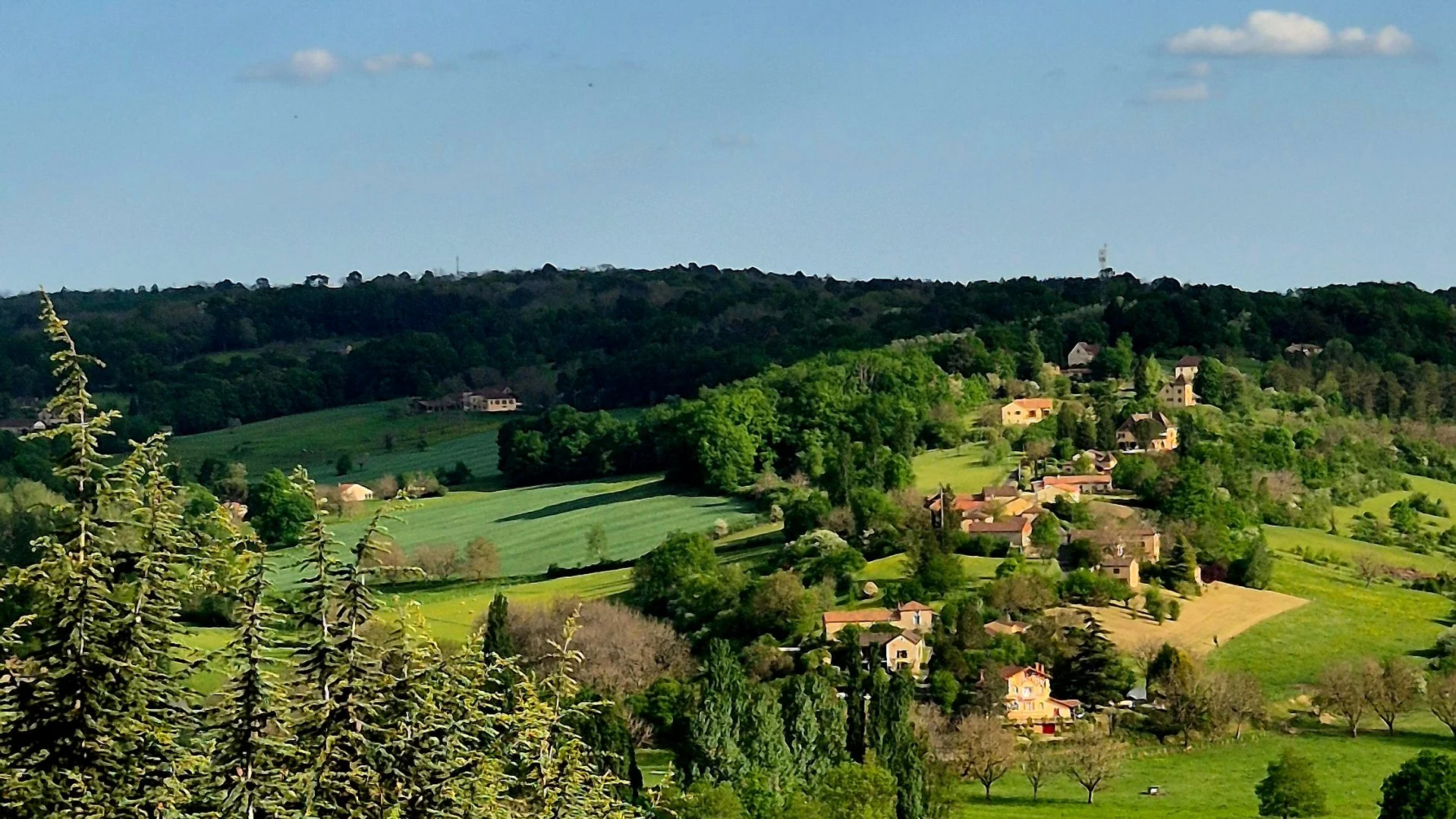 A picture of a view from belvesA scenic landscape of rolling green hills with scattered houses and trees under a blue sky with some clouds.