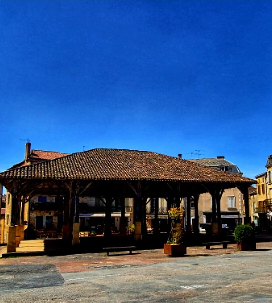 A wooden pavilion with a tiled roof in a town square, surrounded by buildings and planters, under a clear blue sky.