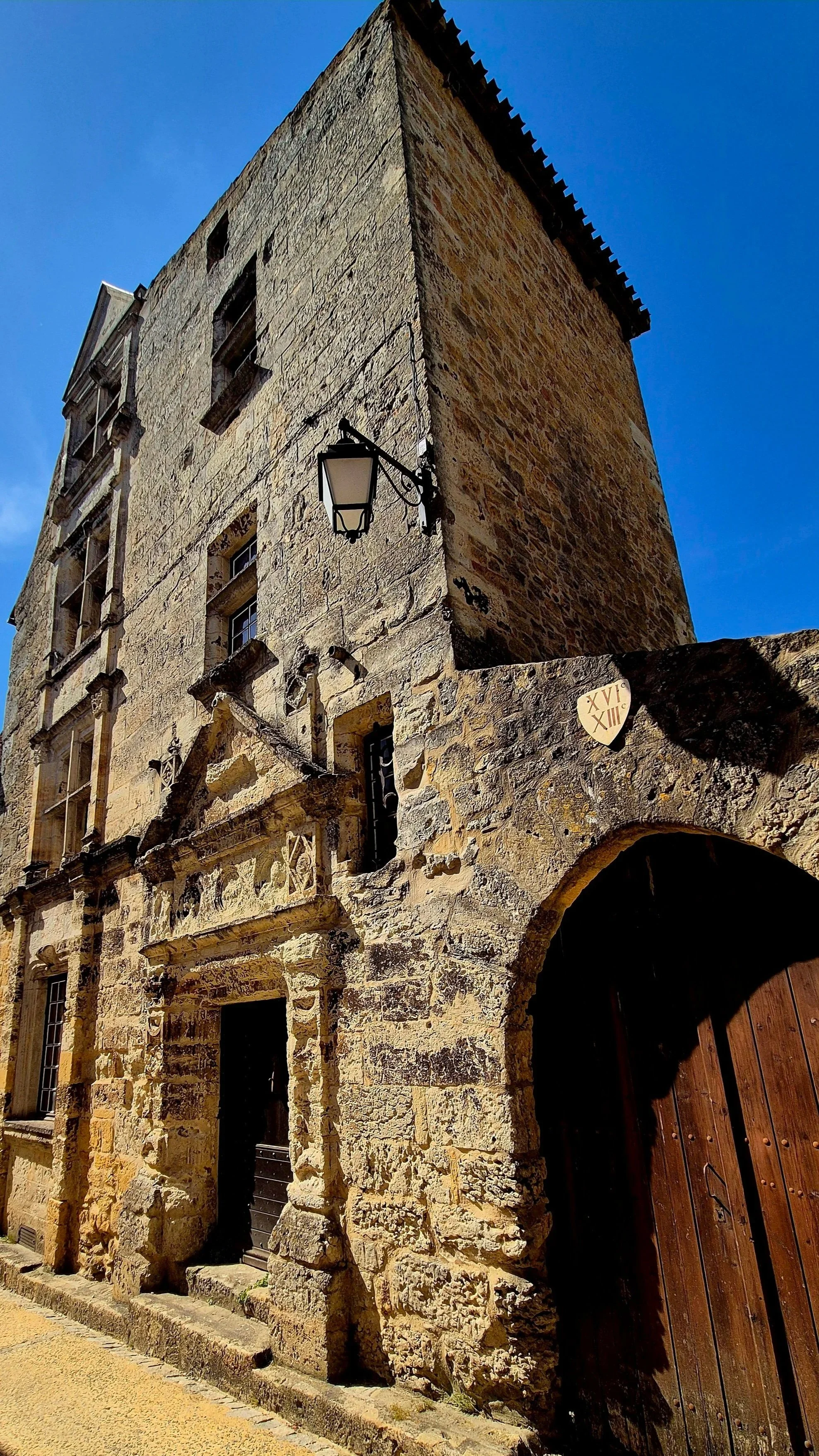 An old stone building with a curved wooden door and lattice windows, a street lamp attached to the corner, a Roman numeral plaque, and a partly ruined facade against a clear blue sky.
