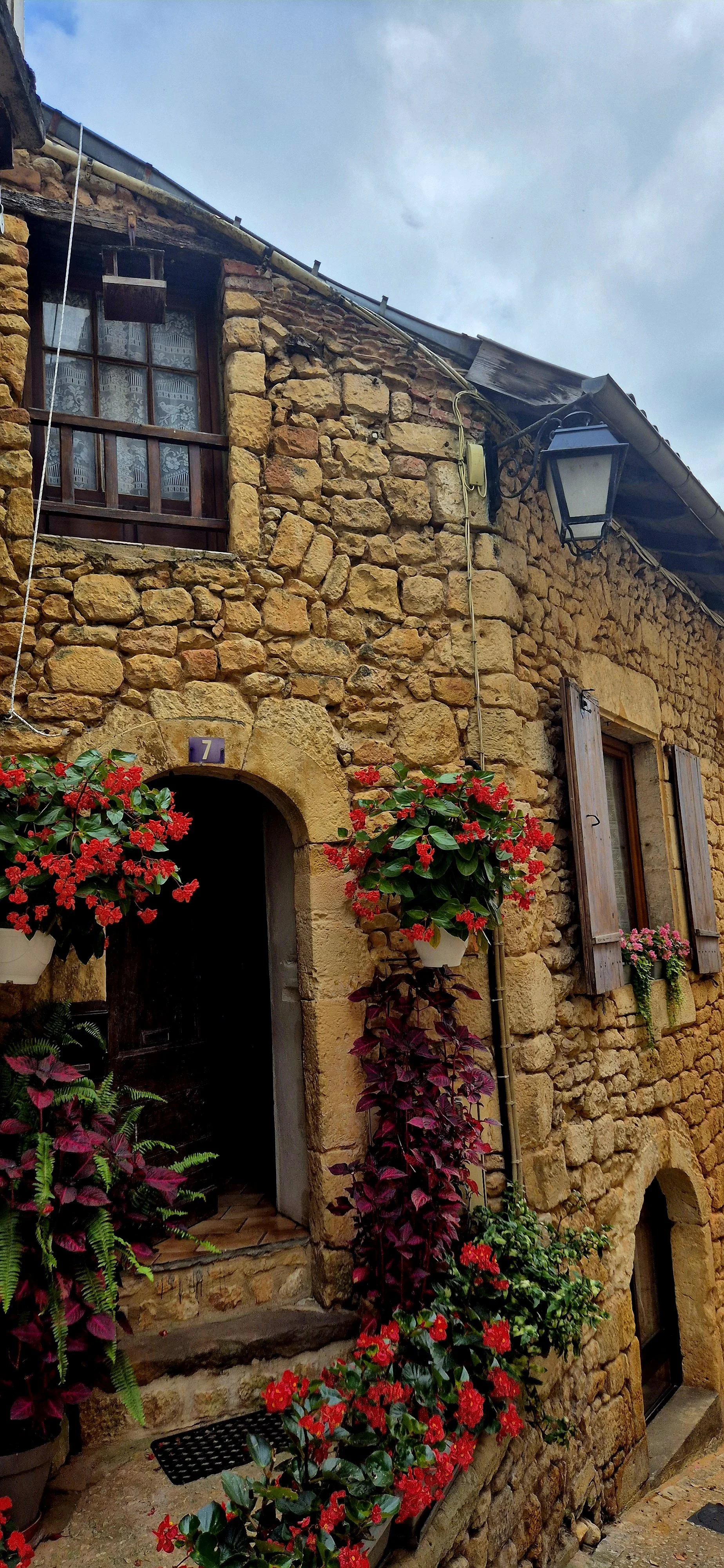 A rustic stone house with wooden shutters, a front door archway, and flower pots with red, pink, and purple flowers, including geraniums and begonias, by the entrance and window.