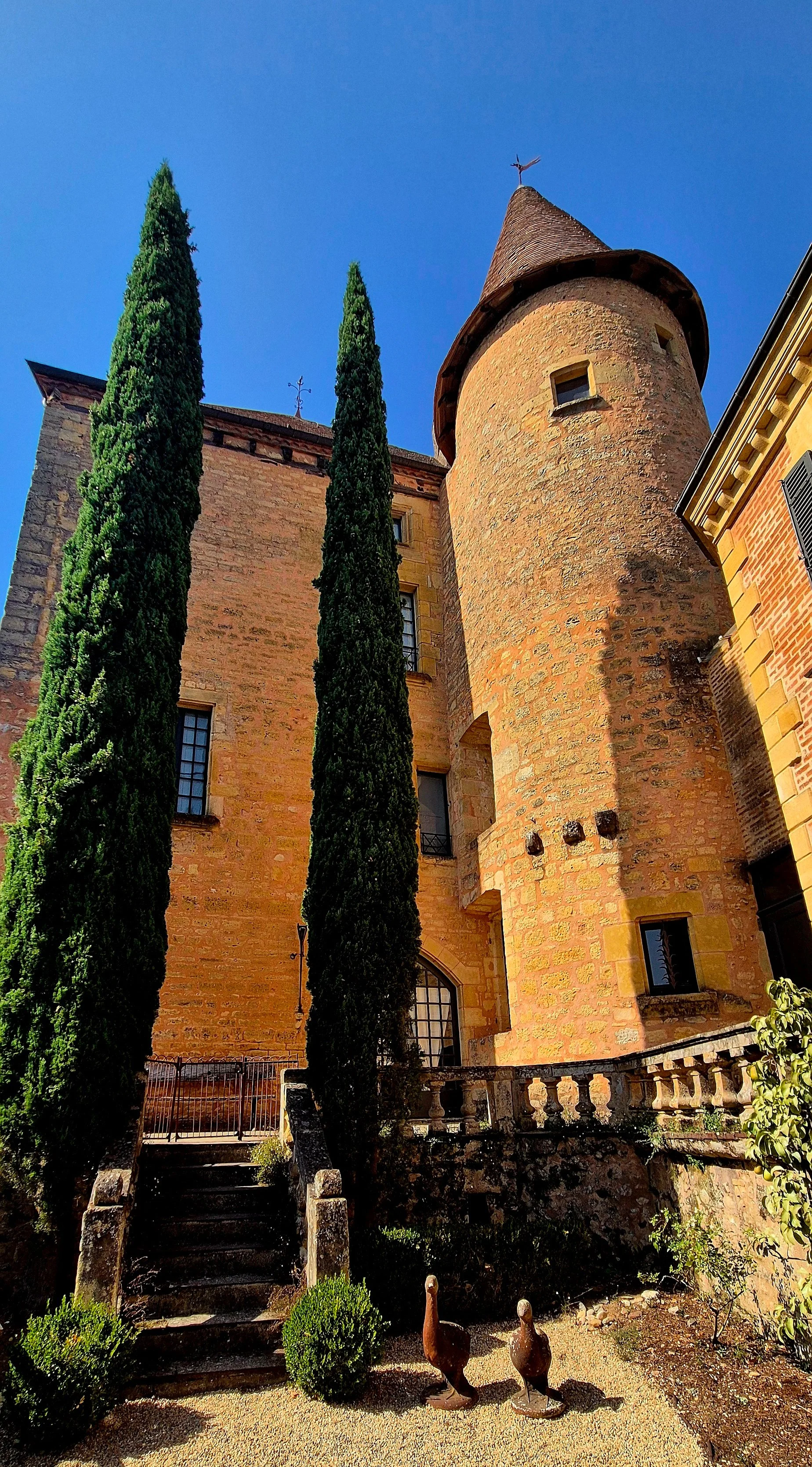 A historic castle with a round tower, stone walls, and tall cypress trees in front. There are stairs leading up to the entrance and two duck statues on the ground.