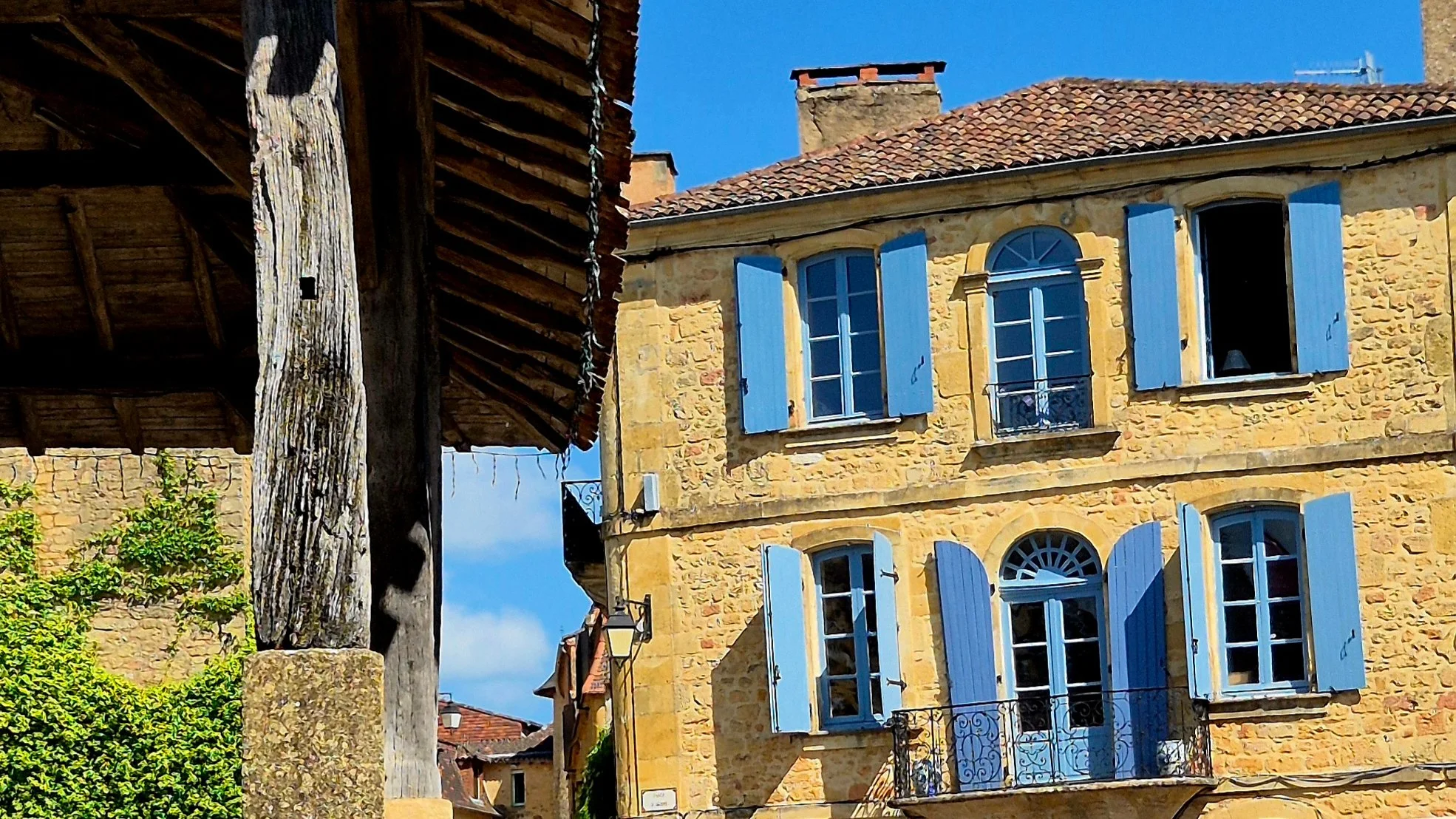 Belves Market Hall in the foreground with a beautiful stone house with sky blue sgutters 
