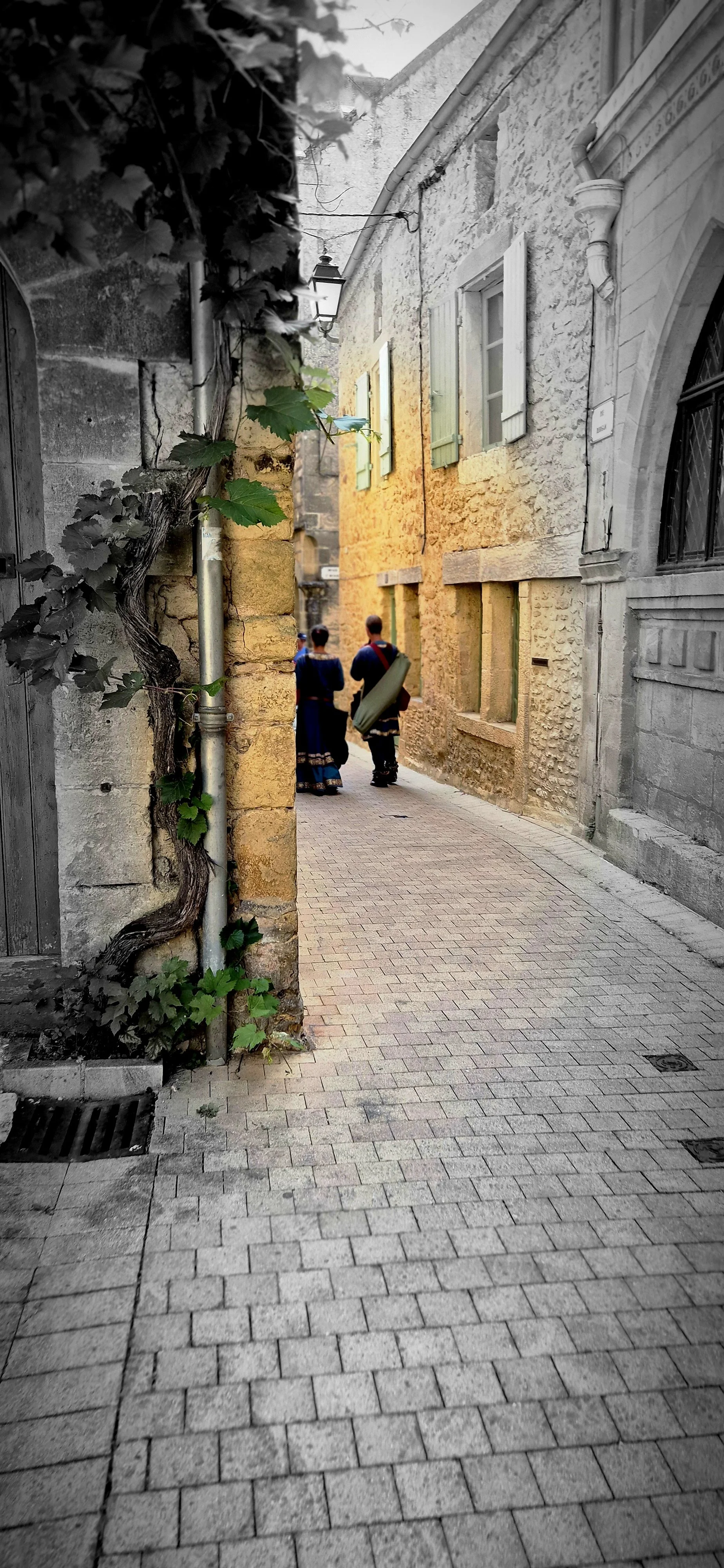 Two people dressed in traditional clothing walking down a narrow cobblestone alley with old stone and brick buildings on either side, some greenery on the left.