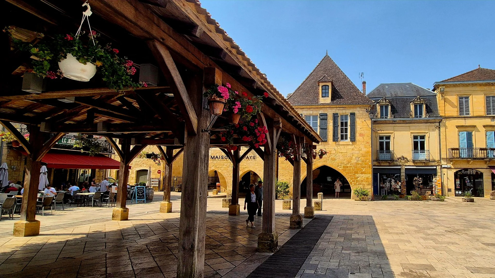 picture of the halle in beumont du perigord with flowers hanging off the roof trusses. Behind are some beautiful shops and houses on the main village square