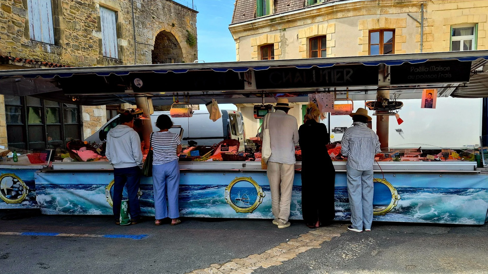 picture of the fish stanll at belves market with customers buying fish. You can also see the arch to Belves castrum in the background