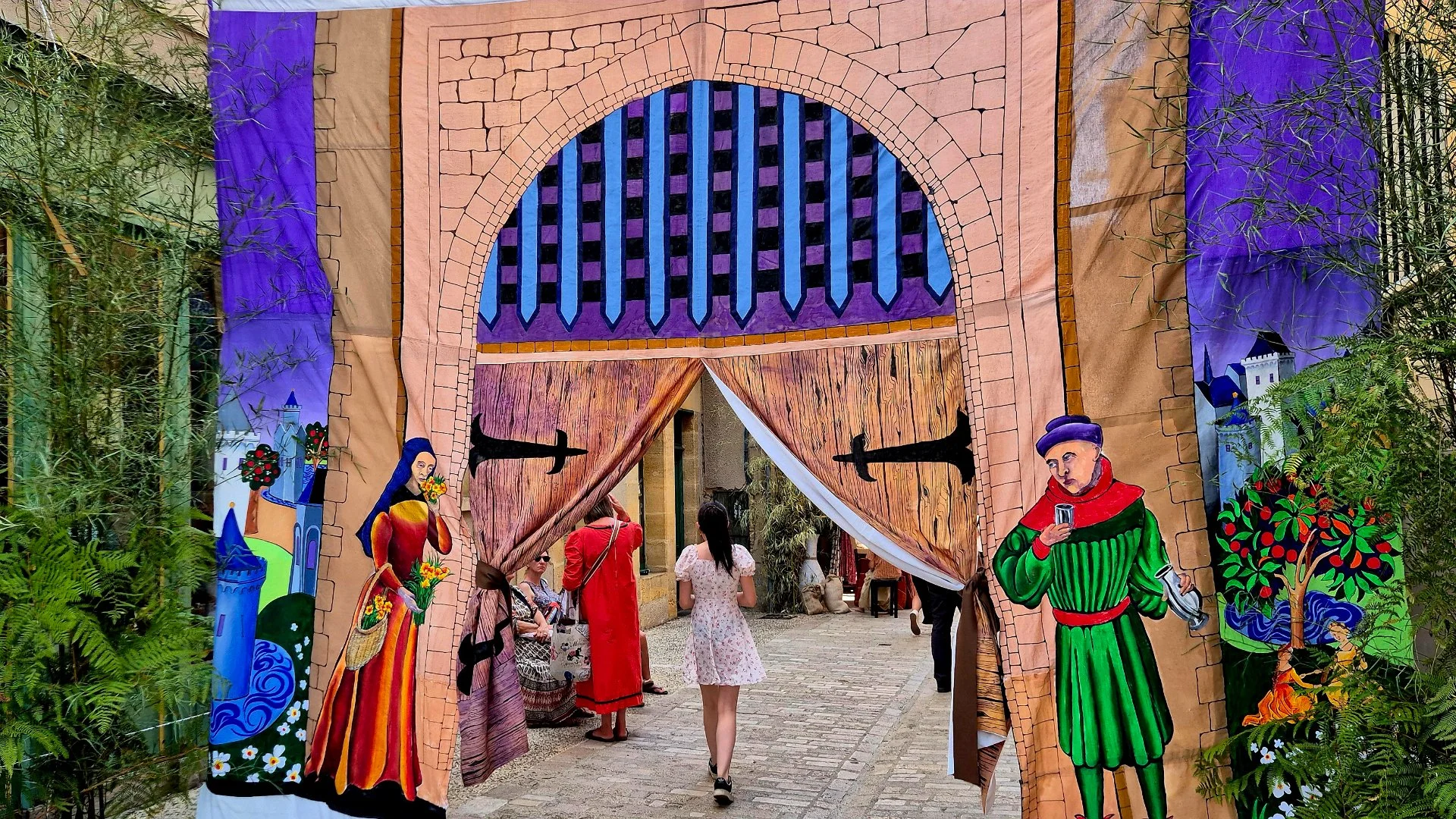 Picture of the Medieval Day in Belves. Colorful medieval-themed archway with painted characters and castle scenes, surrounded by greenery, at an outdoor event.
