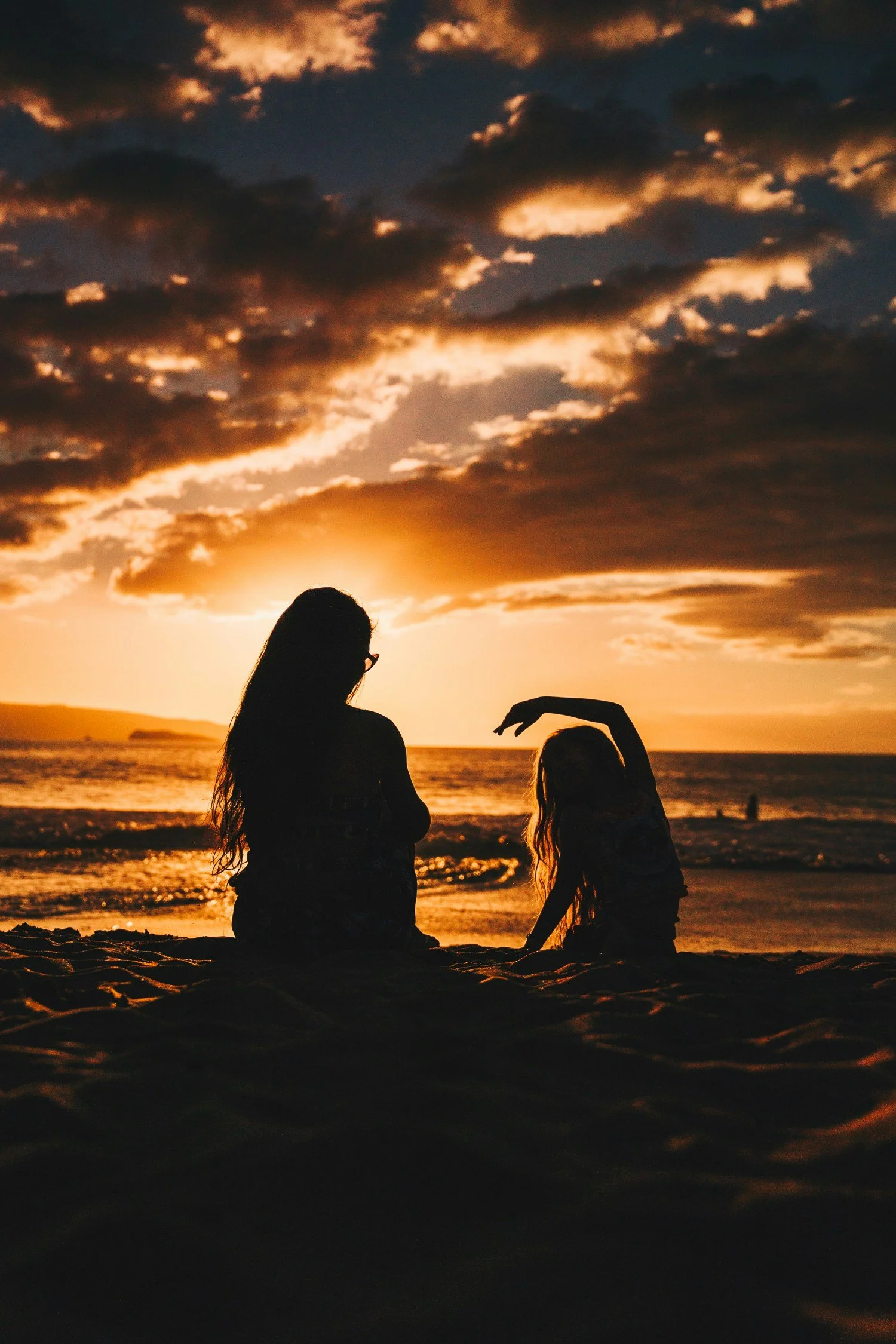 Silhouettes of a woman and a girl sitting on the beach during sunset, with waves and clouds in the sky.
