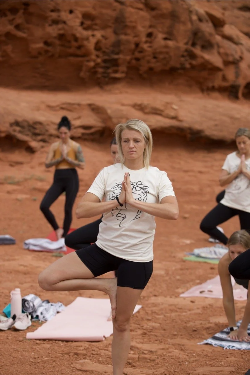 A group of women practicing yoga outdoors on a red dirt surface with a rocky, desert-like background. The woman in the foreground is standing in a tree pose with her hands pressed together in prayer, wearing a white T-shirt with a graphic and black shorts.