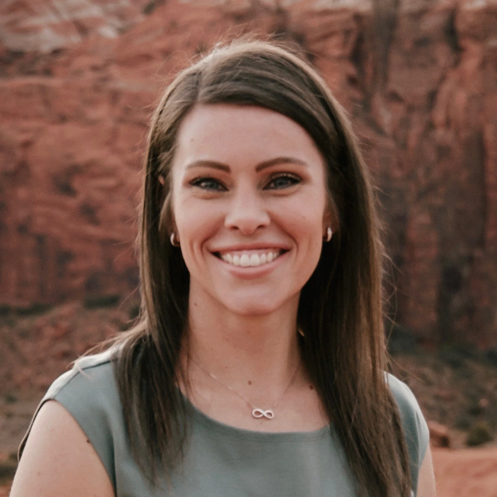 A smiling woman with long brown hair standing outdoors with red rock formations in the background.