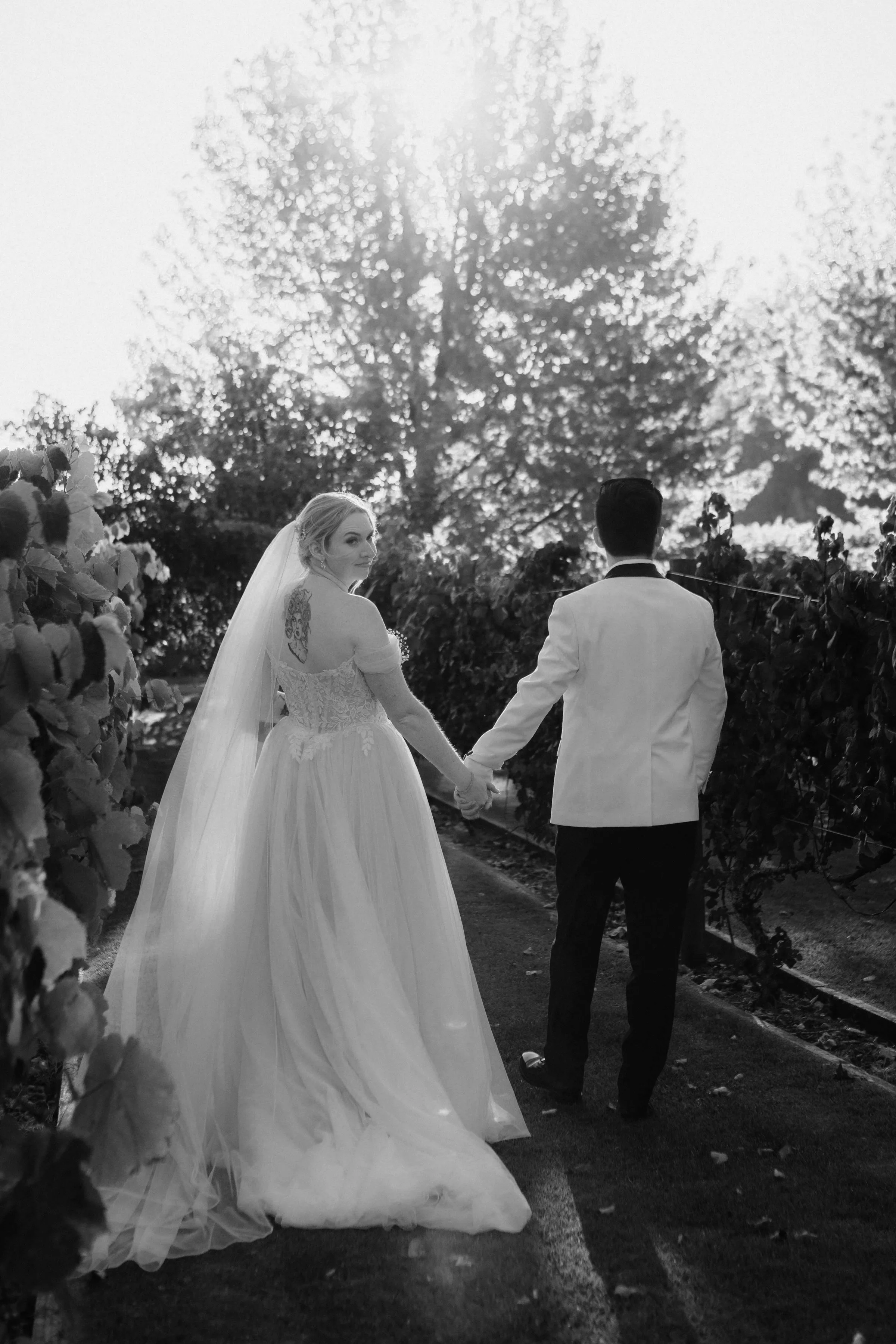 Black and white photo of a bride and groom holding hands on a pathway through a vineyard or garden, with trees in the background, at sunset.