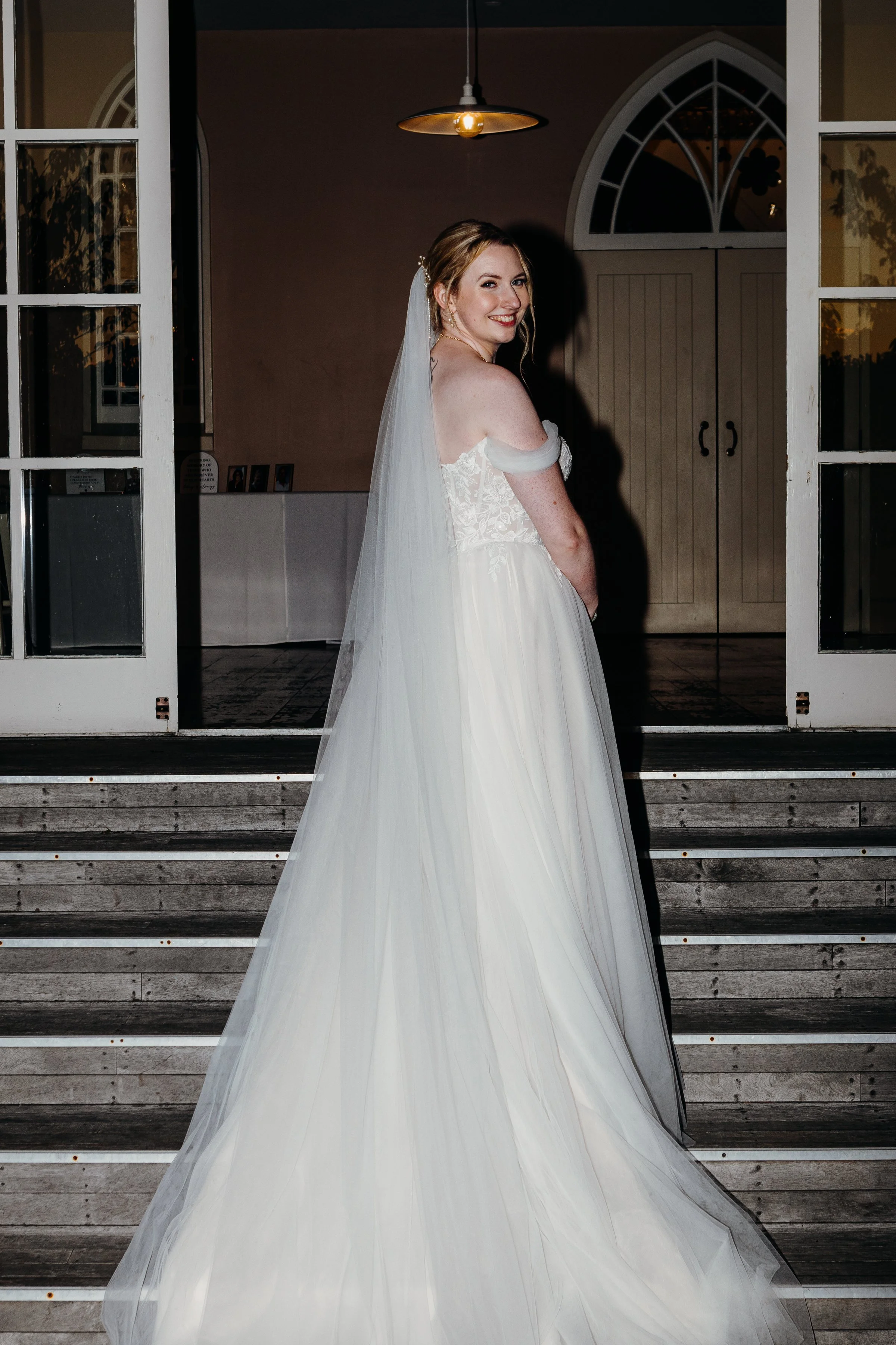 A bride in a white wedding gown and veil standing on wooden steps outside a building, smiling and looking over her shoulder.