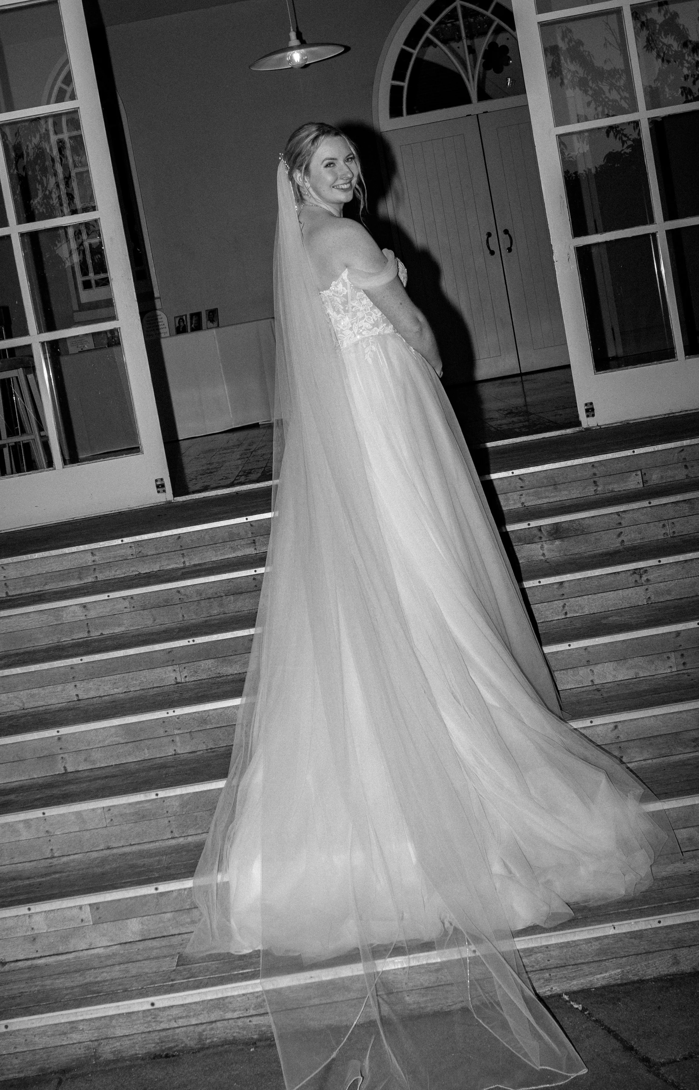 A bride in a wedding dress with a long veil, standing on a staircase inside a building, smiling at the camera.