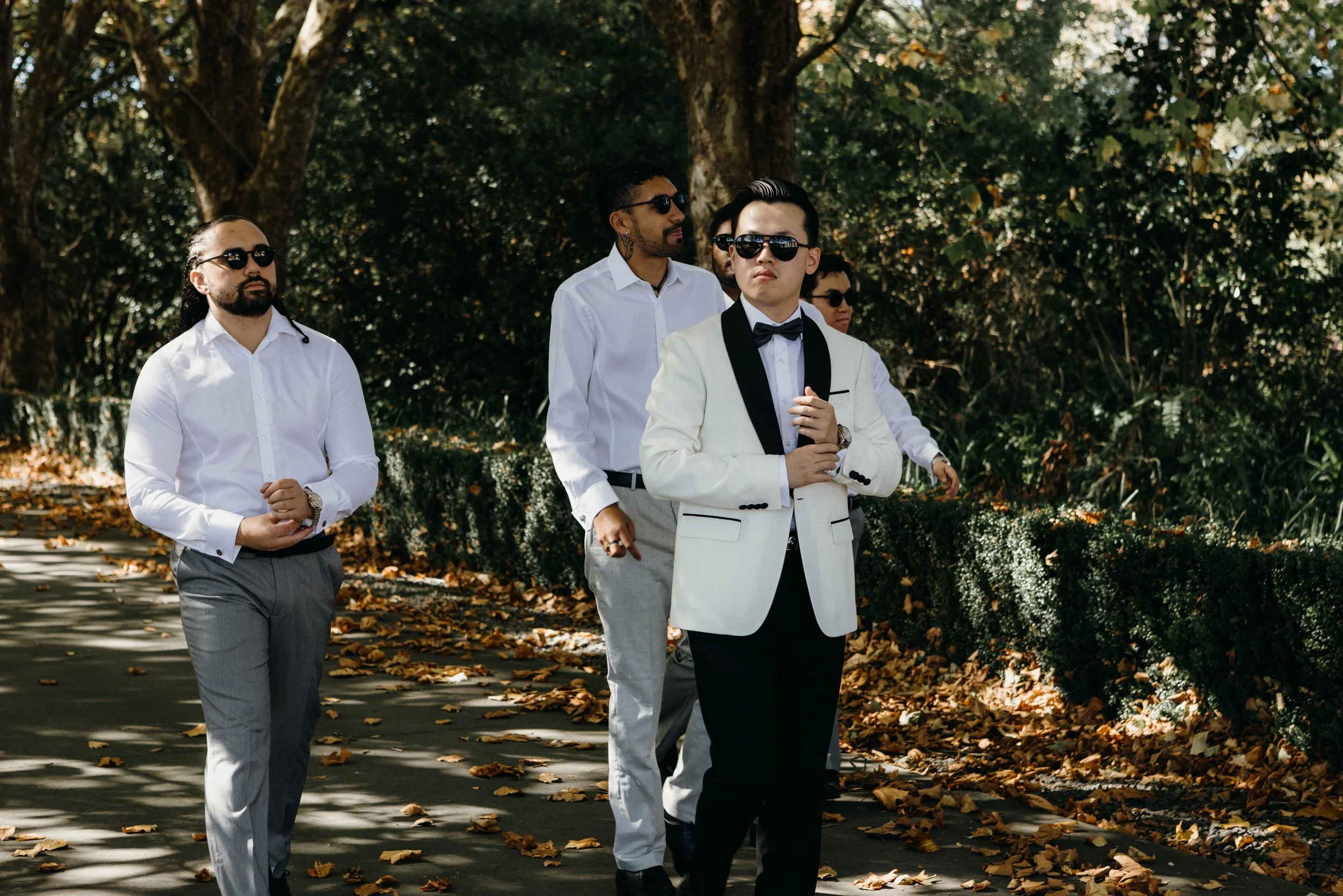 Group of well-dressed men walking outdoors on a sunny day, with trees and fallen leaves in the background.