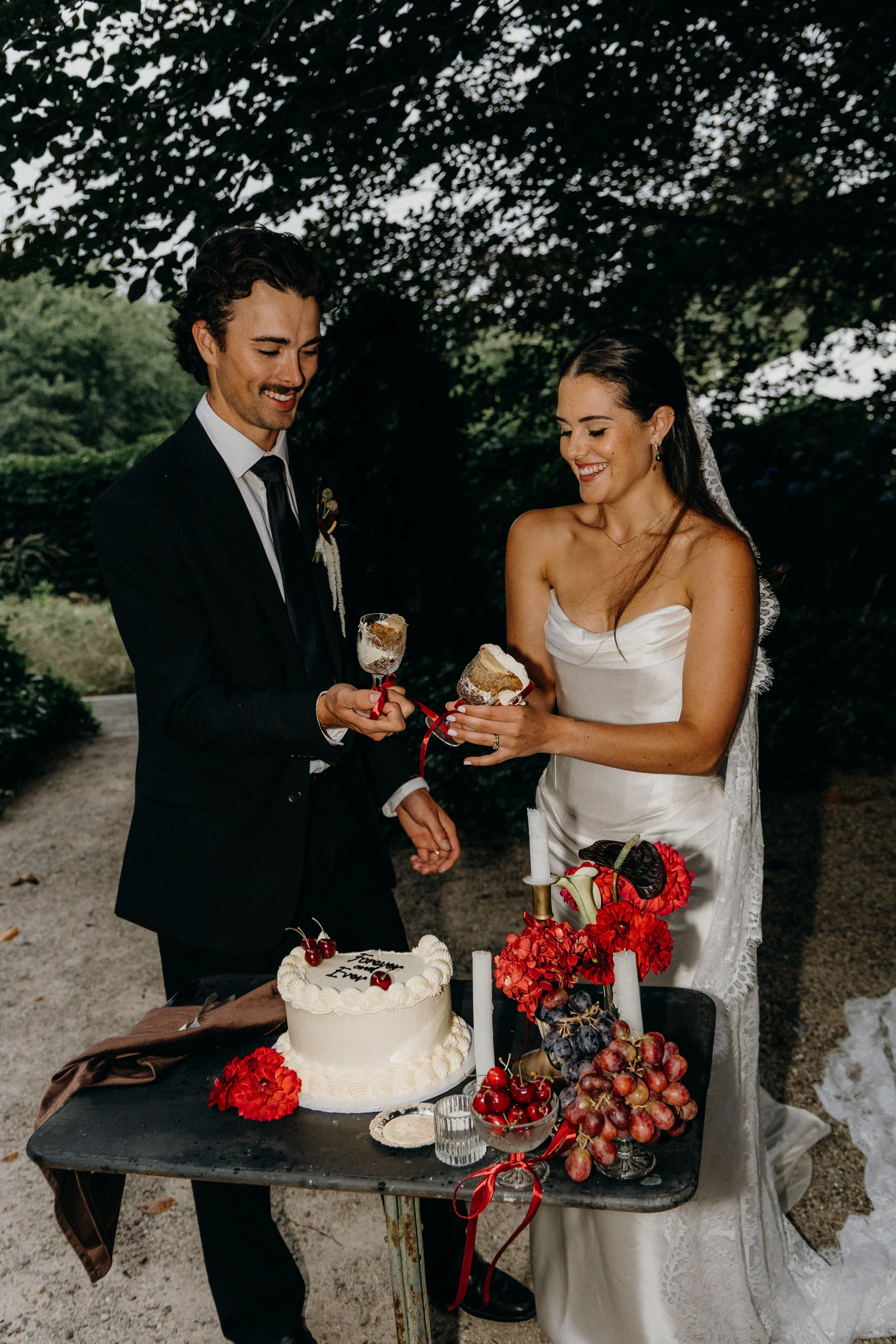 Bride and groom in wedding attire sharing cake outdoors, with wedding cake and fruit decorations on table.