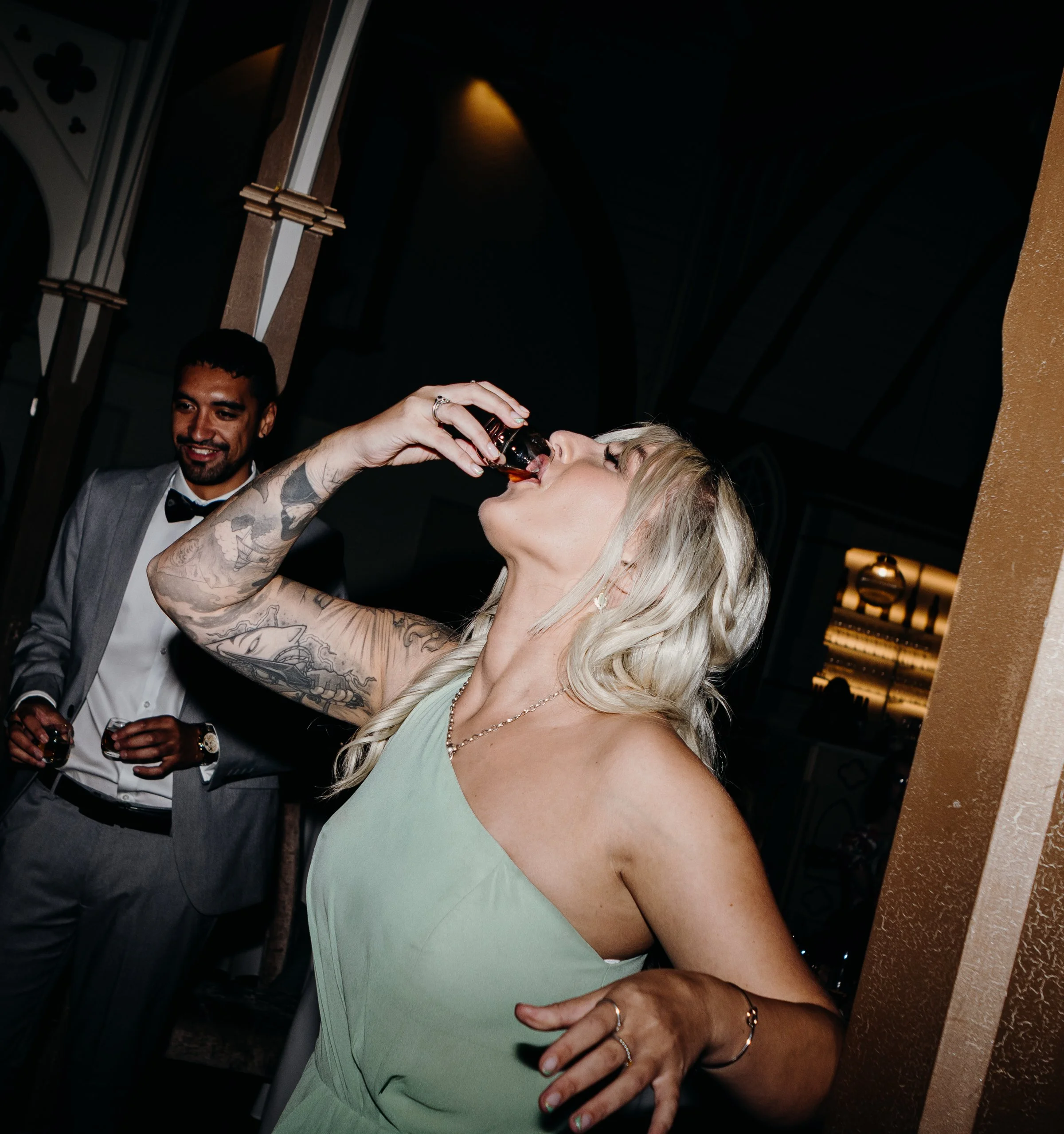 A woman with blonde hair and tattoos on her arm is drinking from a small bottle at a social gathering, while a man in a gray tuxedo looks on smiling.