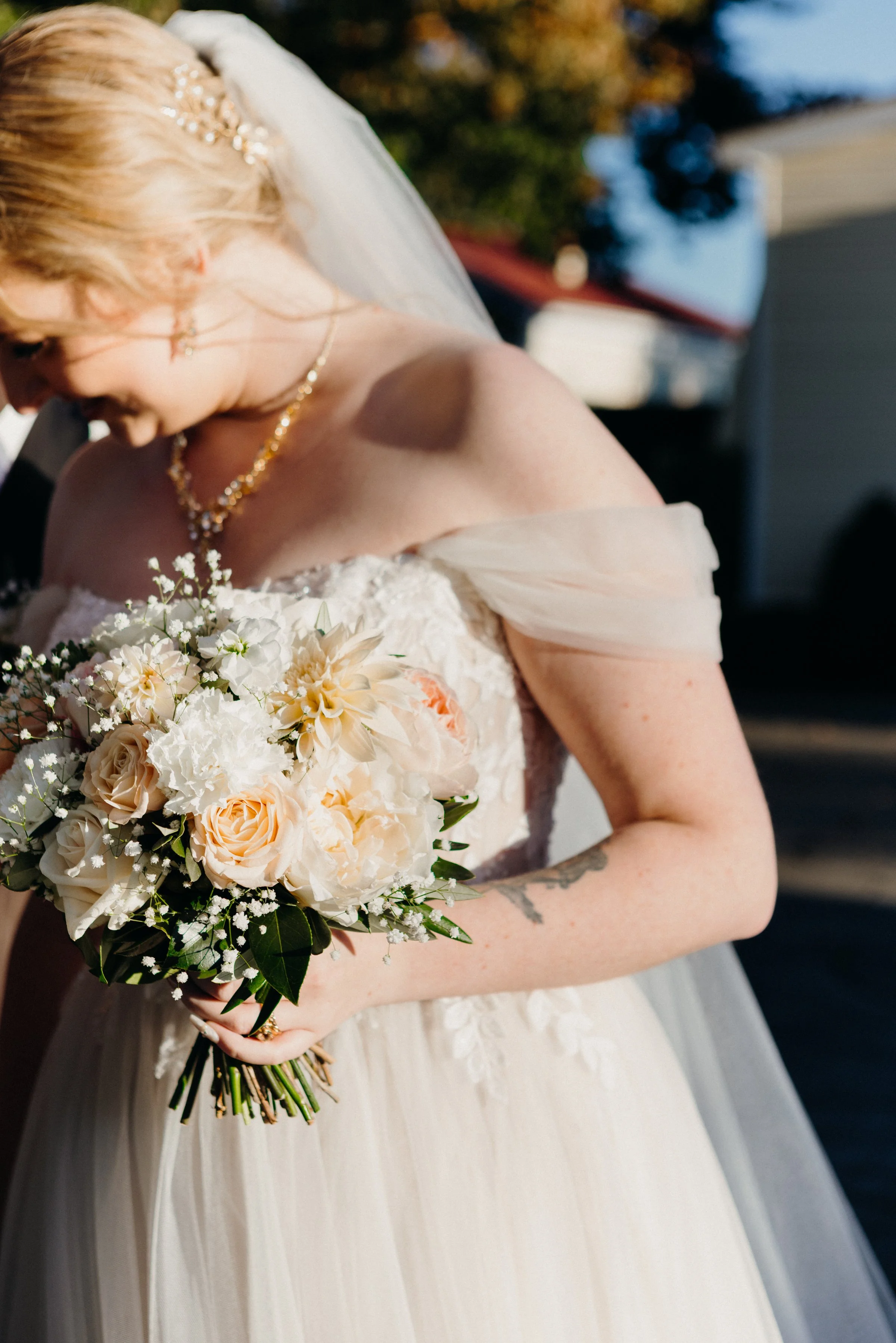 A bride wearing an off-the-shoulder lace wedding dress, holding a bouquet of white, cream, and peach roses and flowers, outdoors during daytime.