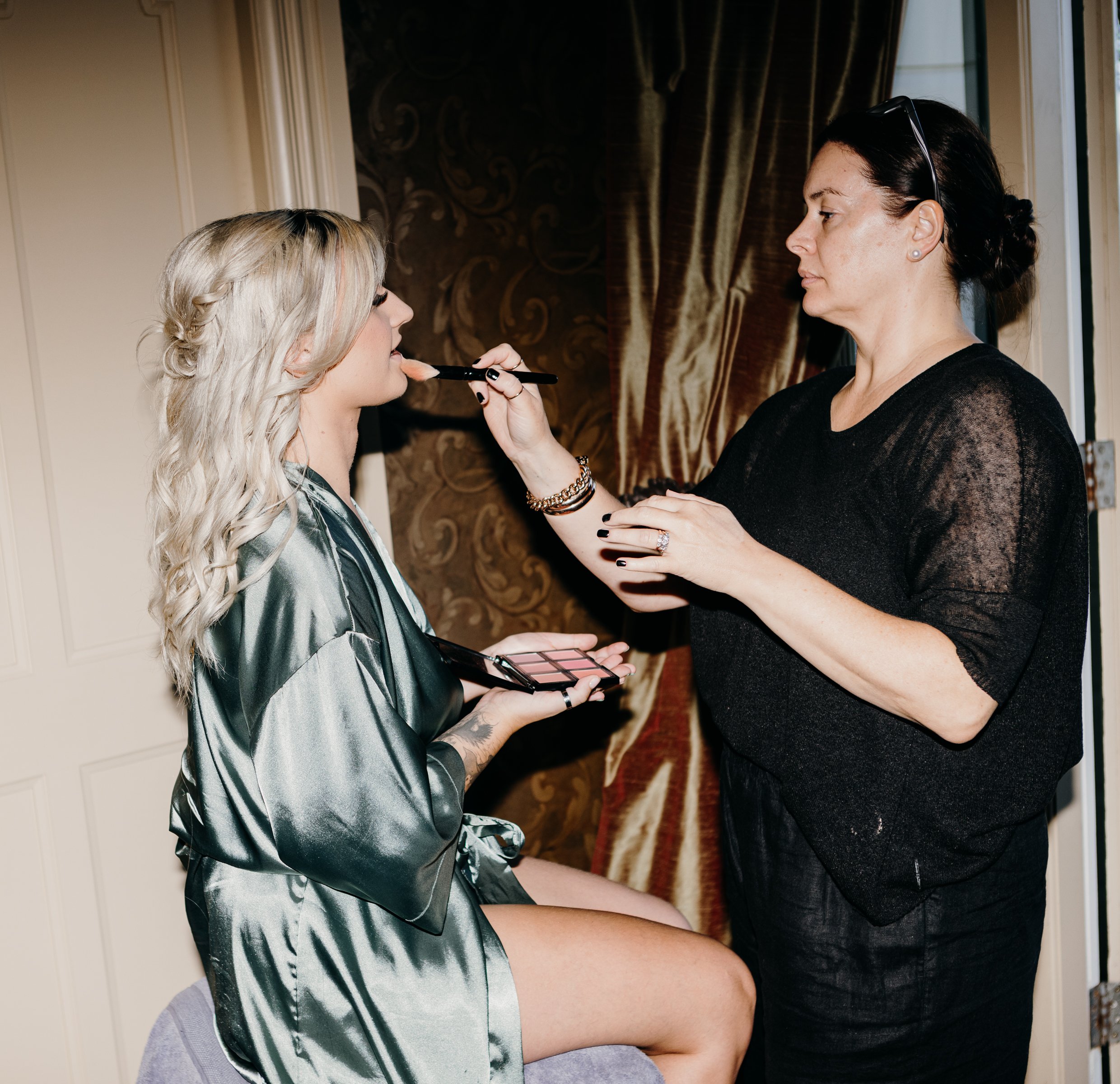 Makeup artist applying makeup to a blonde woman sitting on a chair, with makeup palette in hand, in an interior setting.