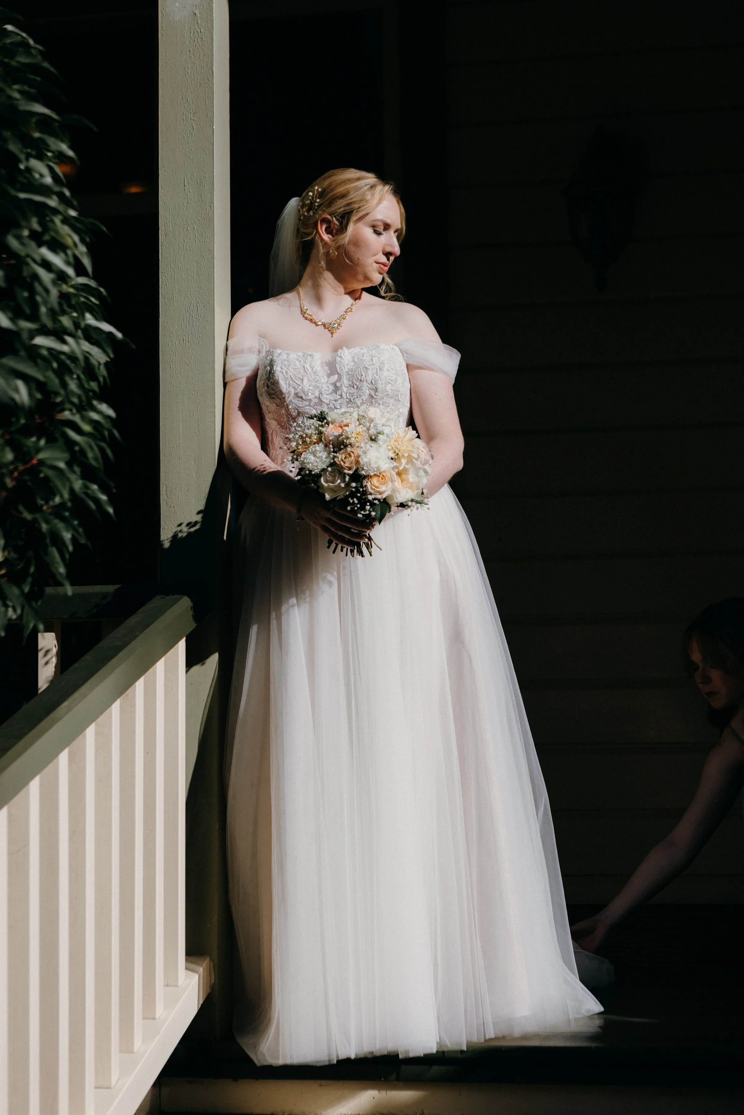 A bride in a white wedding dress holding a bouquet of flowers, standing on a porch with shadows on the wall behind her.