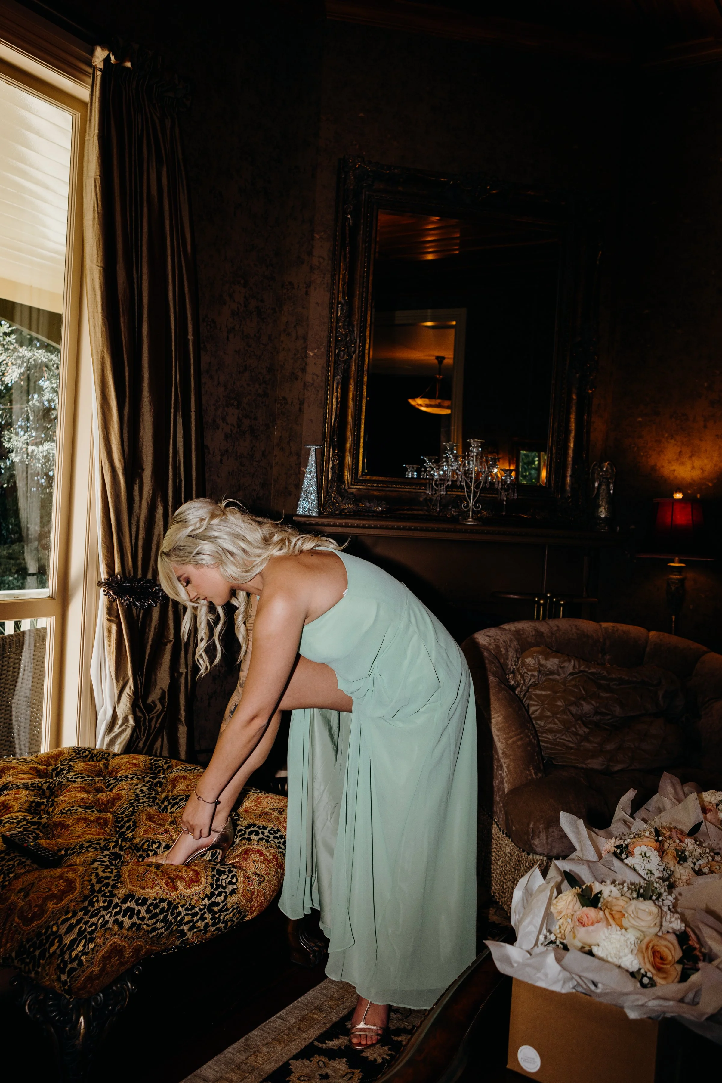 A woman in a light green dress is putting on shoes on a leopard-patterned ottoman in a dimly lit, elegantly decorated room.