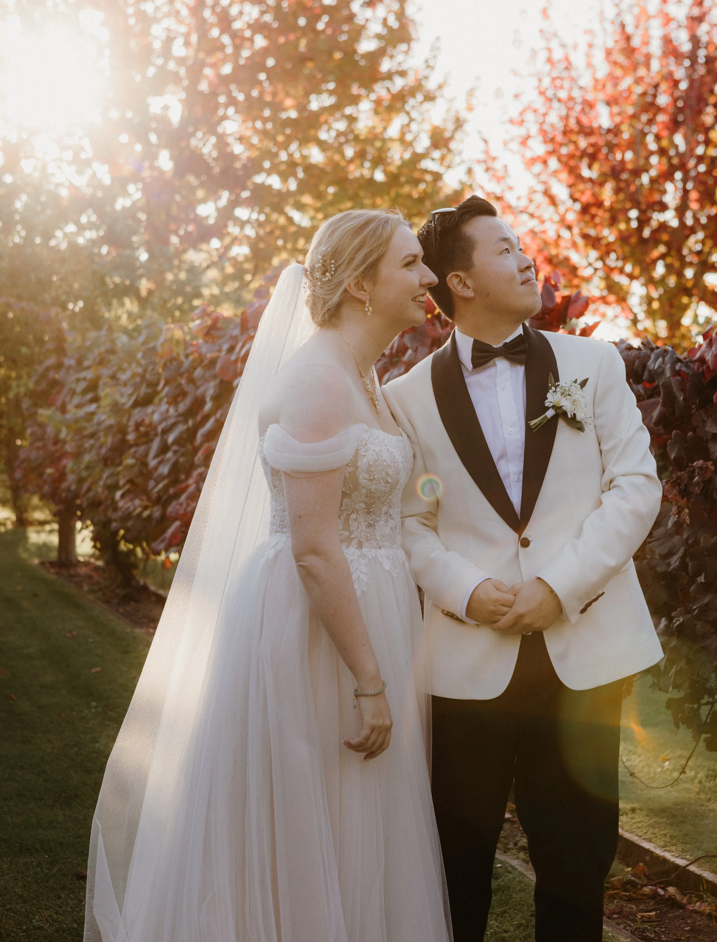 A wedding couple standing outdoors in front of autumn foliage under the sun with a lens flare, the bride in a white wedding dress with veil and jewelry, and the groom in a white tuxedo with black lapels, bow tie, and a boutonniere.