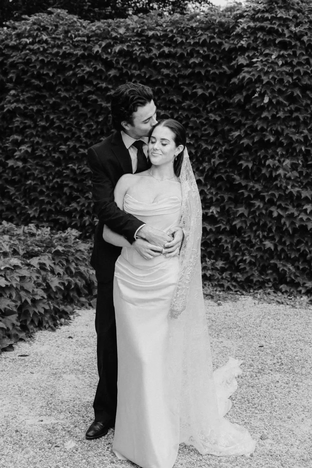 Black and white photo of a bride and groom in formal wedding attire, standing outdoors in front of a hedge. The groom is kissing the bride on the forehead while she smiles with closed eyes, holding her hands in front of her.