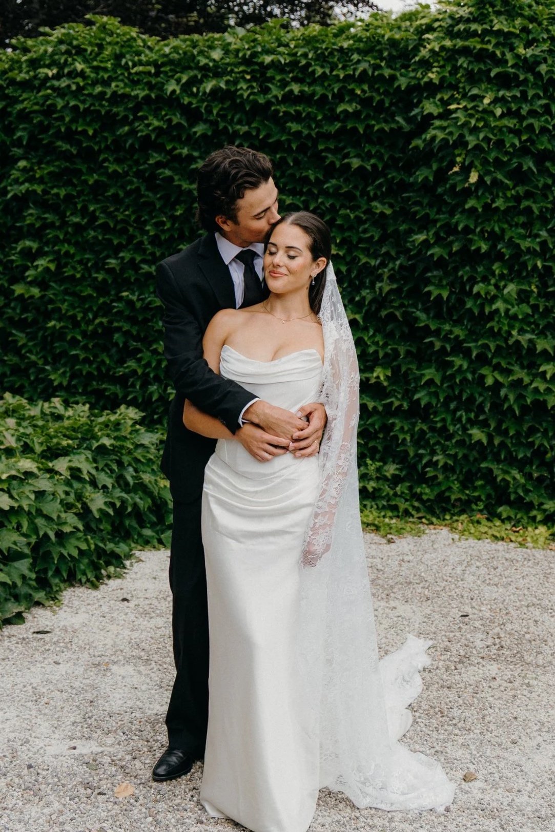 A newlywed couple standing outdoors, with a lush green ivy-covered wall behind them. The groom, dressed in a black suit, gently kisses the bride on the forehead while holding her waist. The bride wears a white strapless wedding gown with a lace veil, and has her eyes closed, smiling softly.
