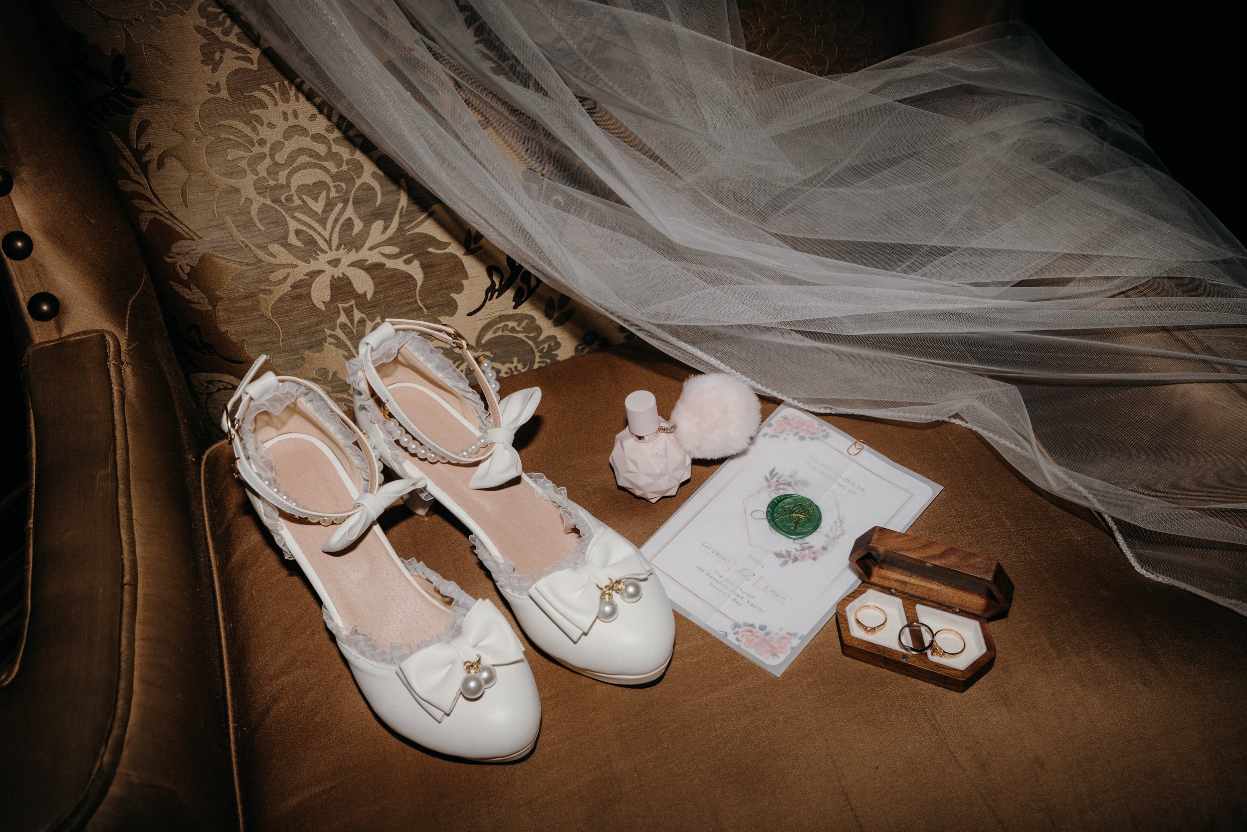 A pair of white wedding shoes with bows and pearl decorations, a pink perfume bottle with a fluffy pom-pom, a wedding invitation, two rings in a wooden box, and a small green stamp on a floral letter.