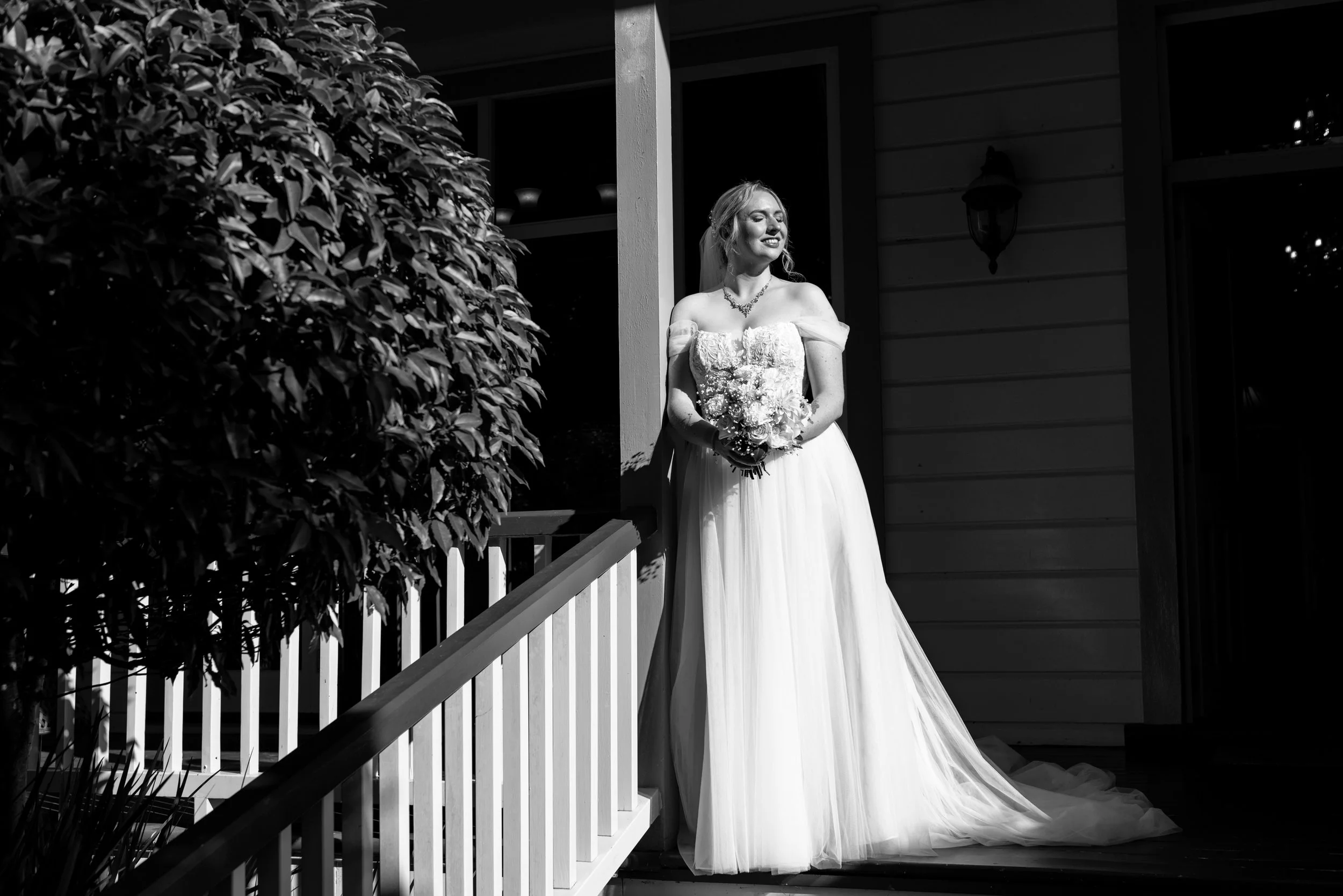 A smiling bride in a wedding dress holding a bouquet, standing on the porch of a house with dark siding, a light fixture, and a railing, next to a bush, with sunlight illuminating her.