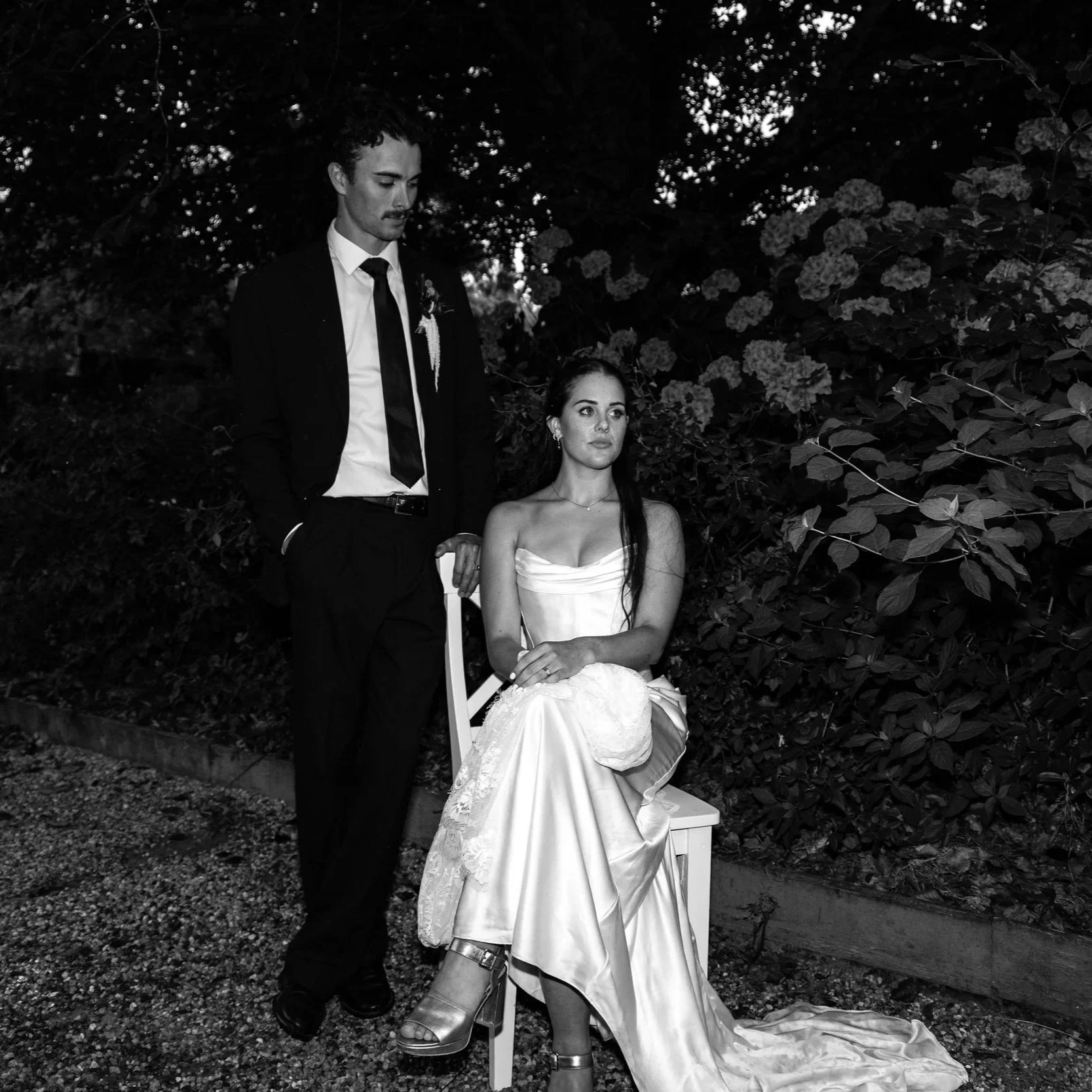 A black and white photo of a man in a suit and a woman in a wedding dress outdoors, with flowers and bushes in the background. The woman is seated on a chair, and the man is standing next to her, looking down.