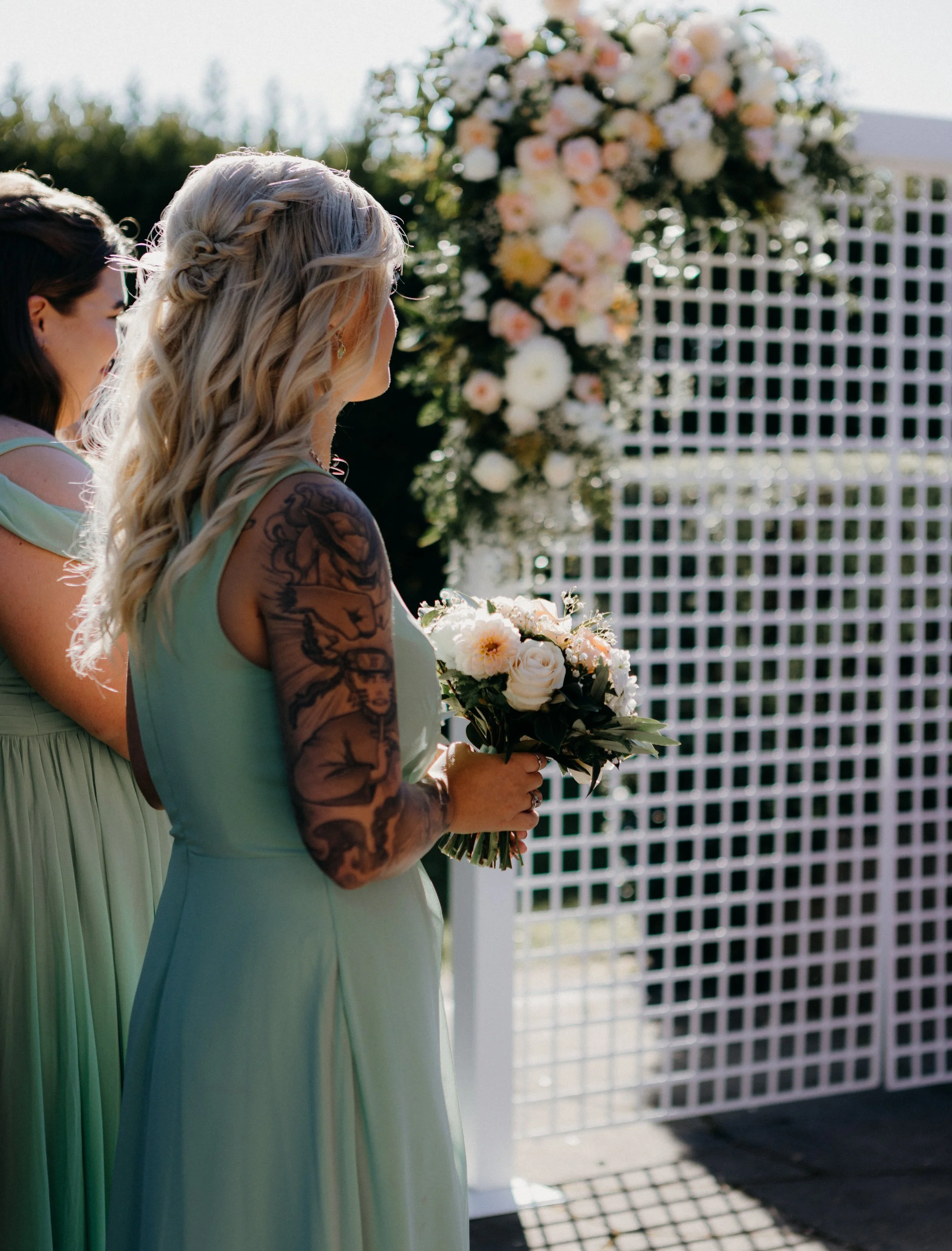 A woman with blonde curly hair and tattoos on her arm holding a bouquet of white roses and flowers at an outdoor wedding or event, with a floral arch and lattice backdrop in the background.