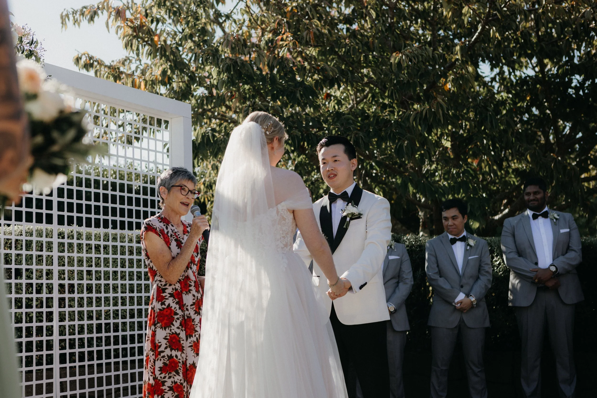 A wedding ceremony outdoors with a bride and groom holding hands, exchanging vows, surrounded by groomsmen in gray suits, an officiant speaking, and green trees in the background.