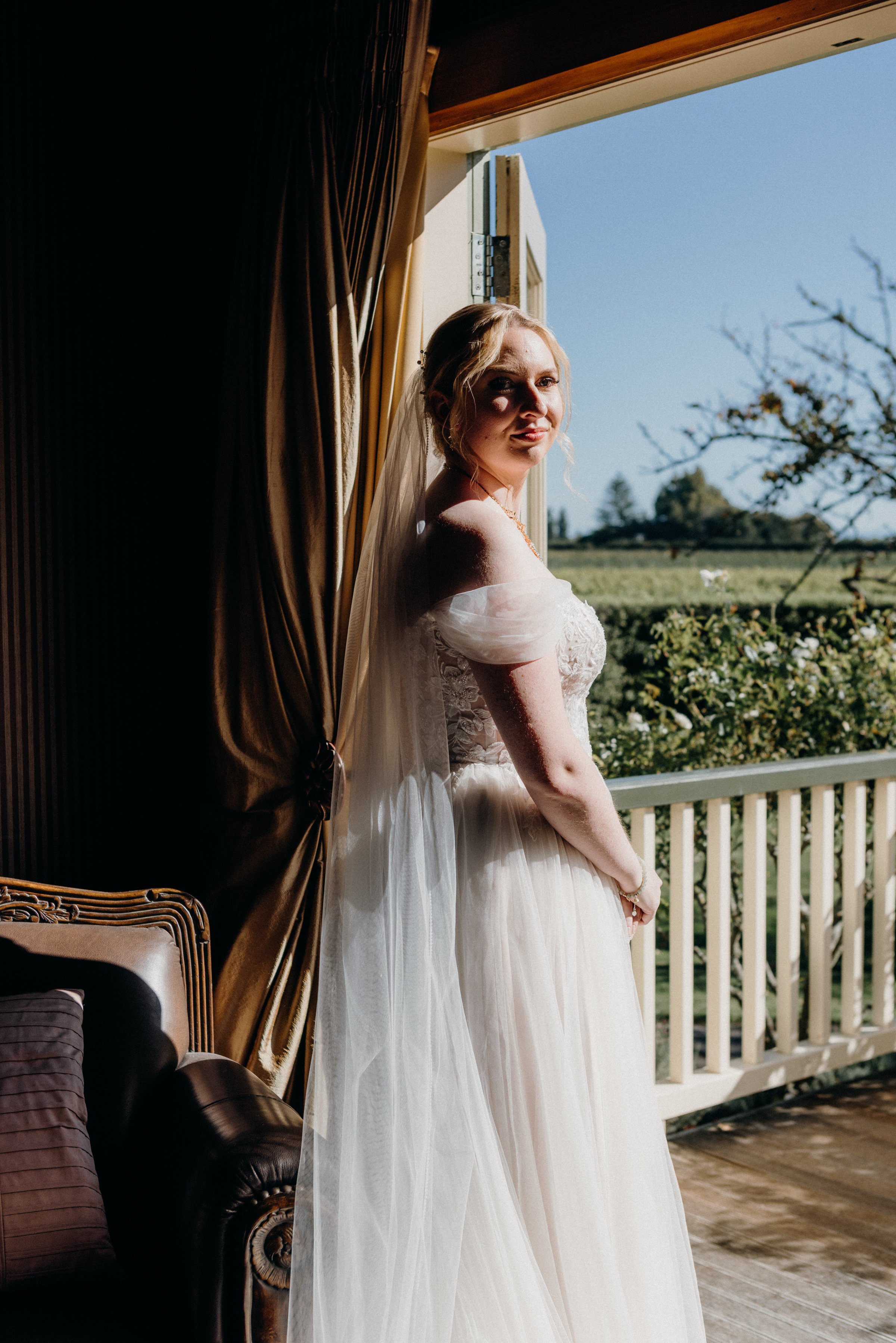 A bride in a wedding dress standing indoors near an open window with sunlight illuminating her, overlooking a green landscape and blue sky outside.