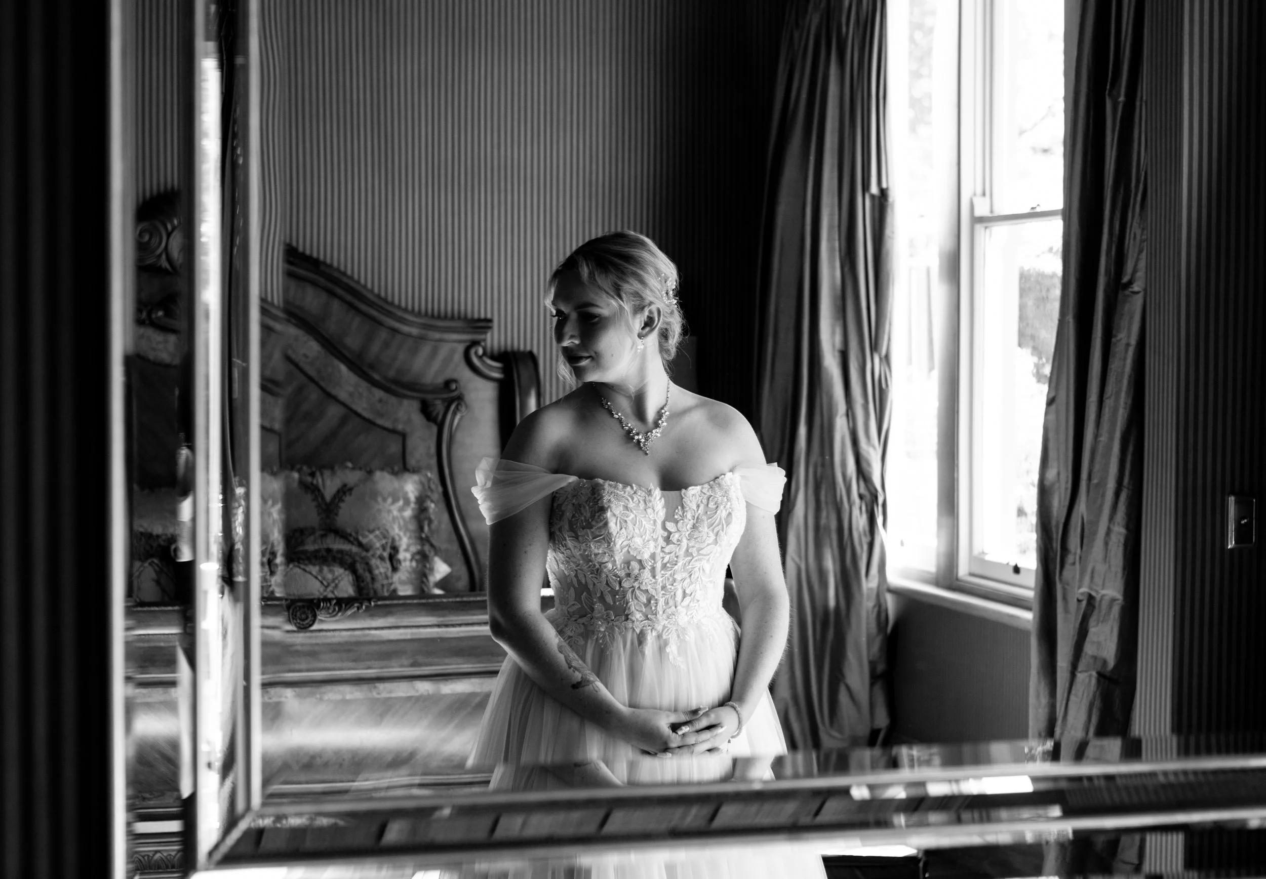 A bride in a wedding dress with off-the-shoulder sleeves standing in a room with a large mirror, window, and curtains, looking to her left.