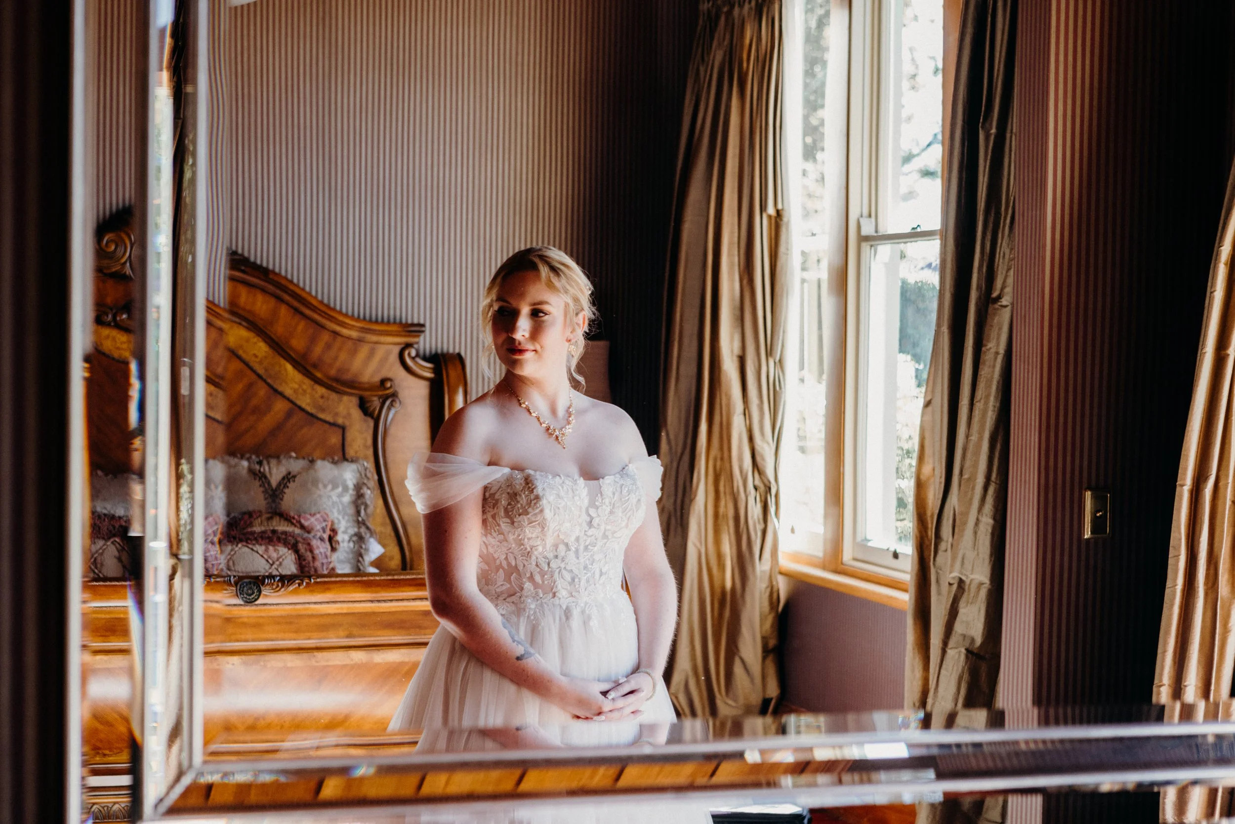 A young woman in a white wedding dress standing in front of a mirror, looking to the side, in a room with wooden furniture and a window with curtains.