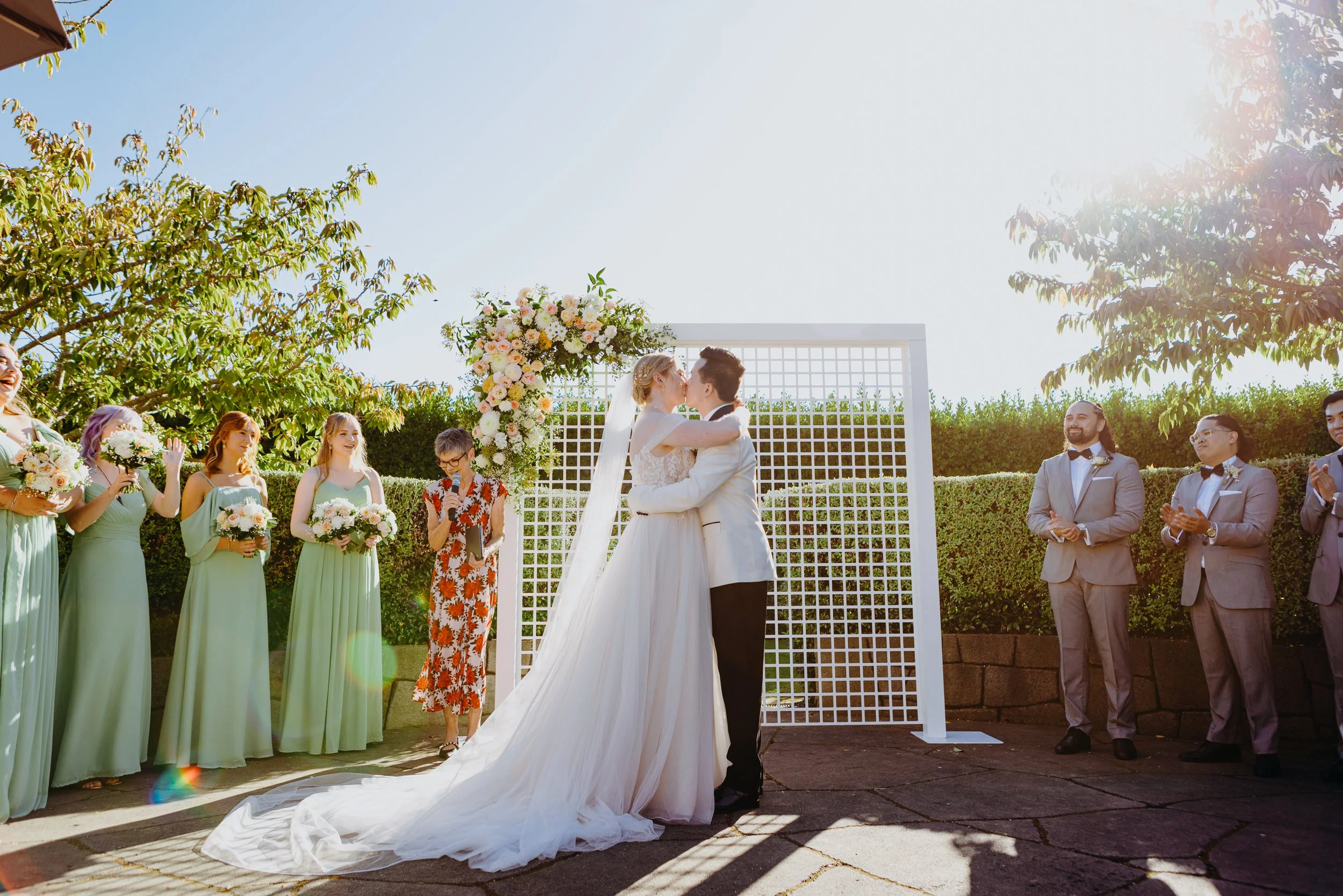 A wedding ceremony outdoors with a bride and groom kissing, surrounded by bridesmaids in green dresses holding bouquets, and groomsmen in suits with bow ties, on a sunny day with trees and a floral arch.