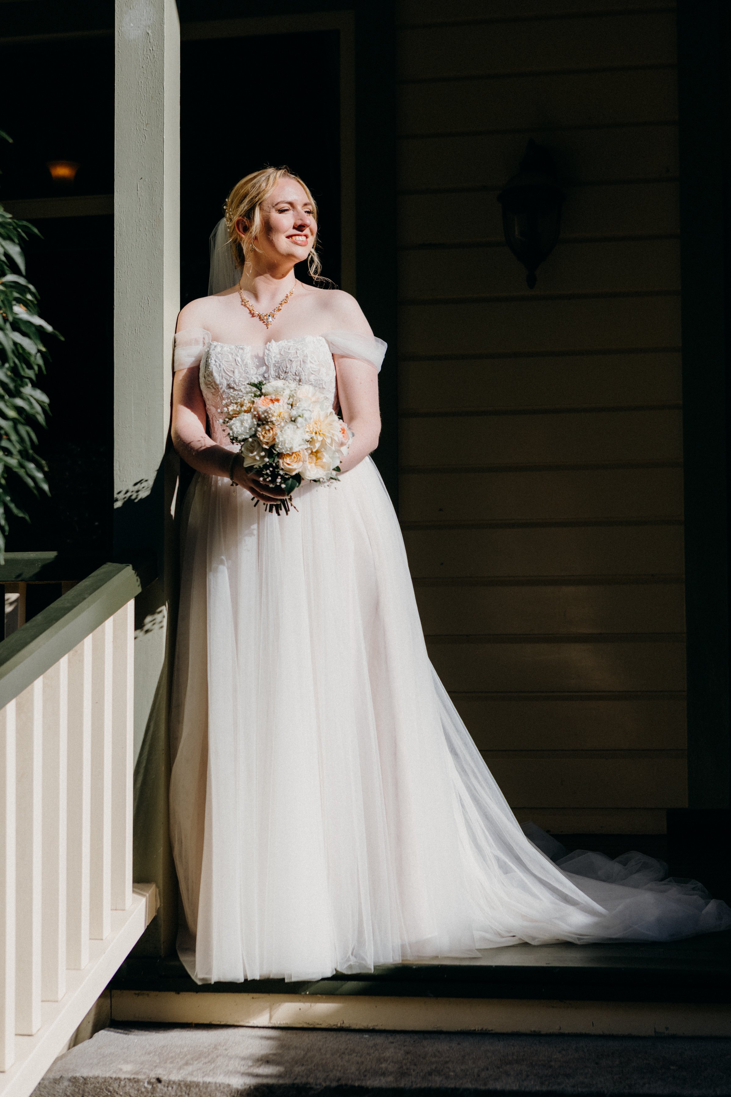 A bride in a white wedding dress holding a bouquet, standing on a porch at night, smiling and looking to the side.