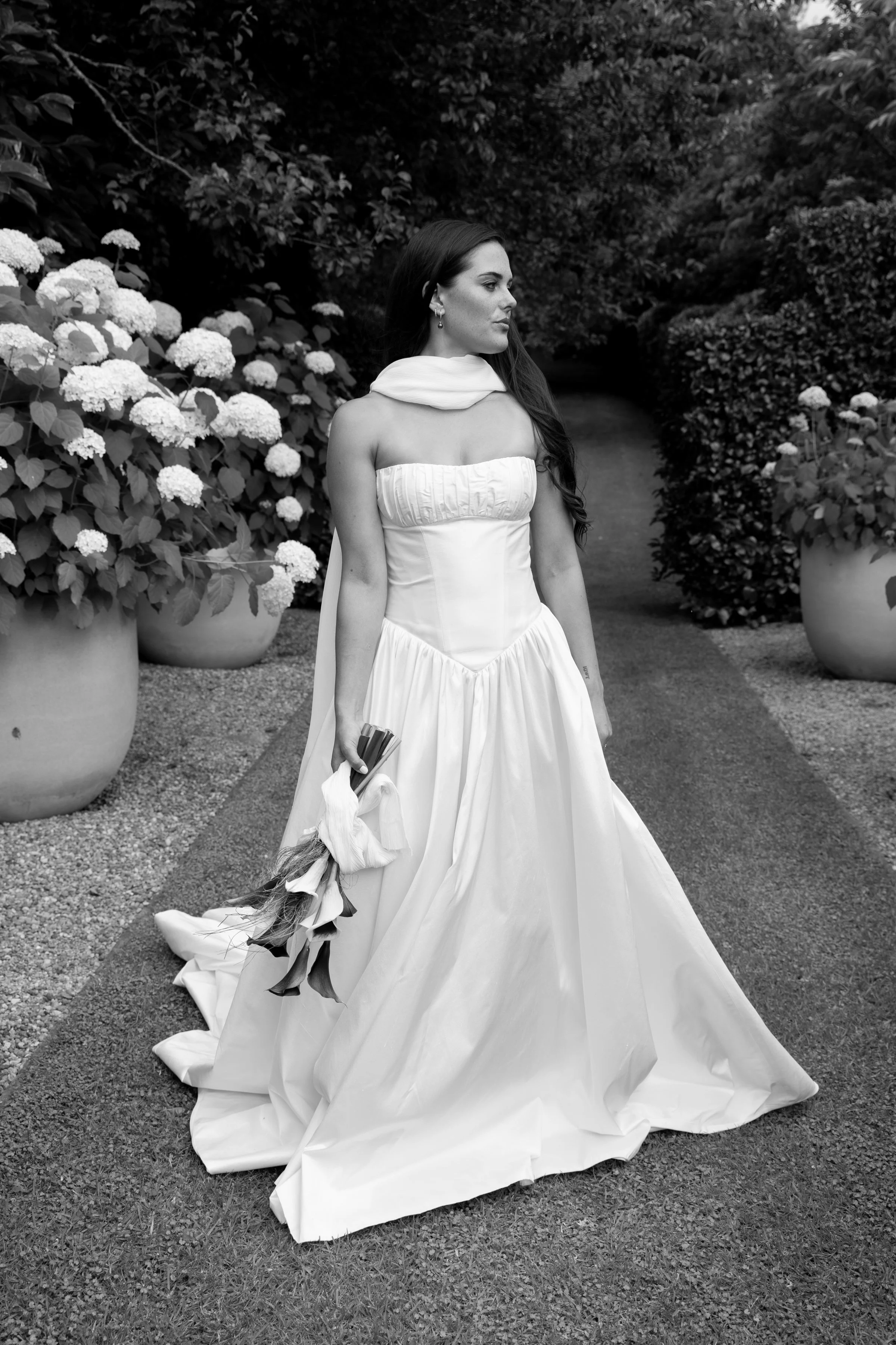 A woman in a wedding dress holding a bouquet, standing on a path surrounded by large potted plants and lush foliage.