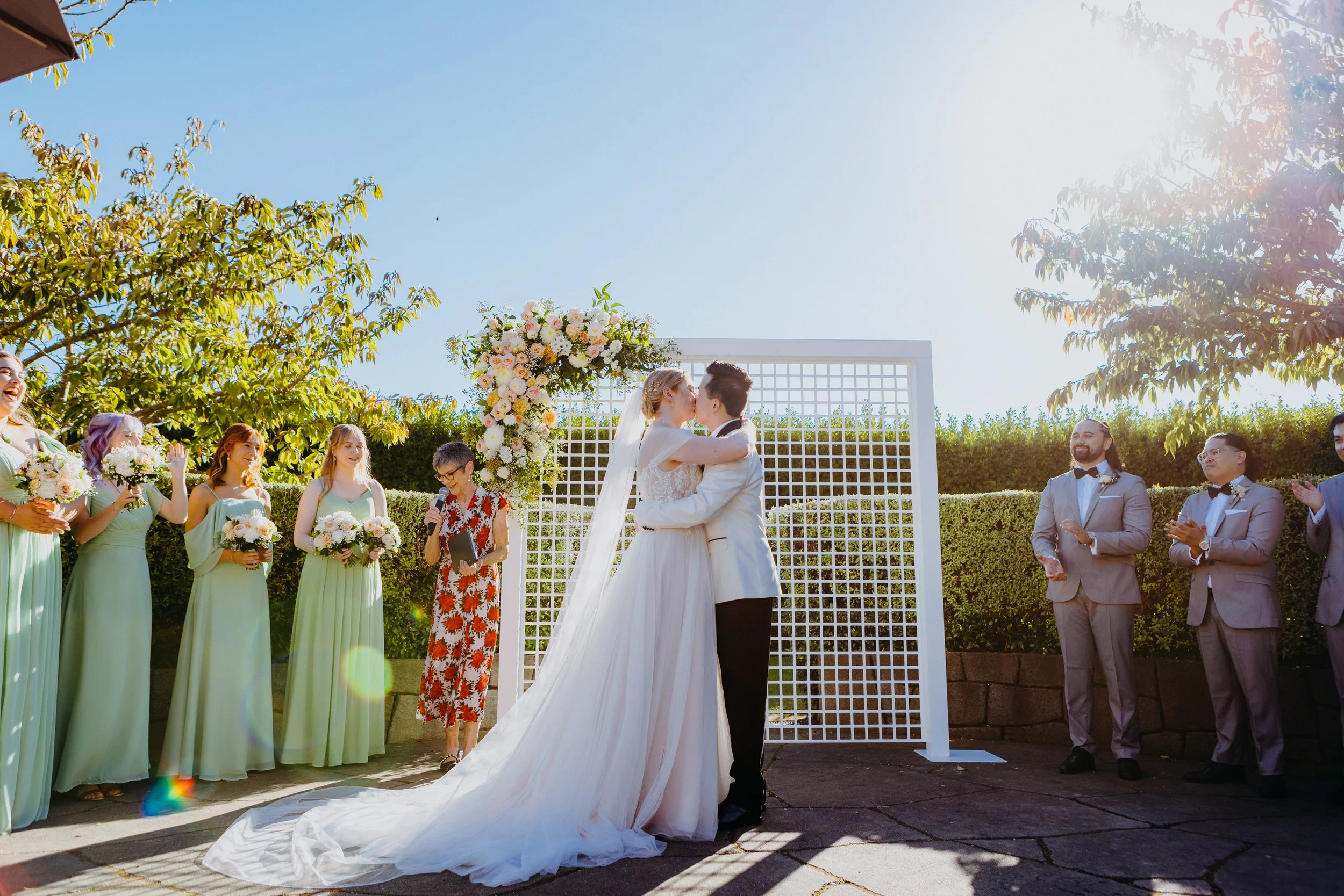 Couple dressed in wedding attire kissing during outdoor wedding ceremony, surrounded by bridesmaids and groomsmen, with officiant holding a microphone and floral arch behind them on sunny day.