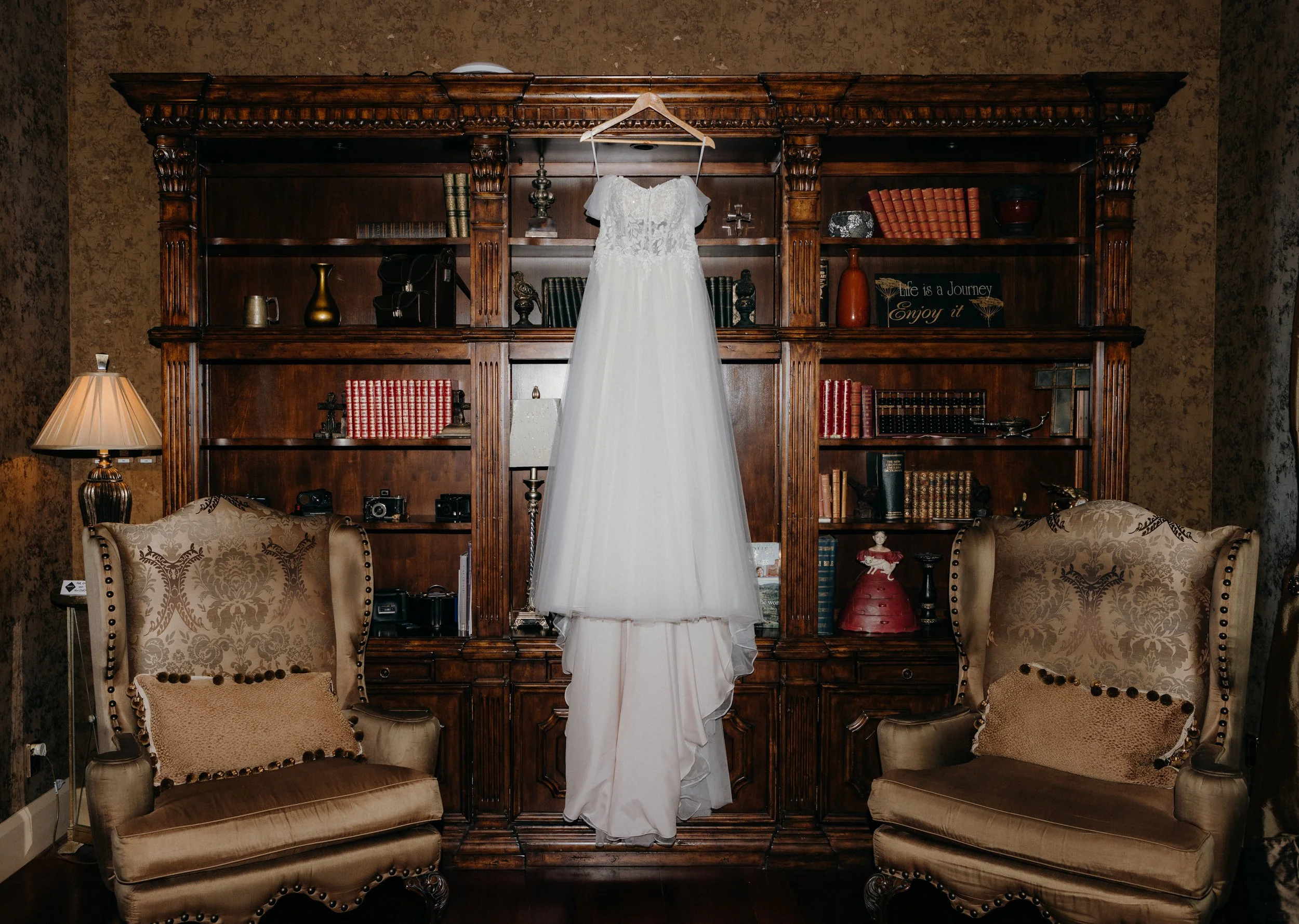 A white wedding dress hanging in front of a large wooden bookshelf with decorative items, two upholstered armchairs with pillows flanking it, and a lamp on the left side.