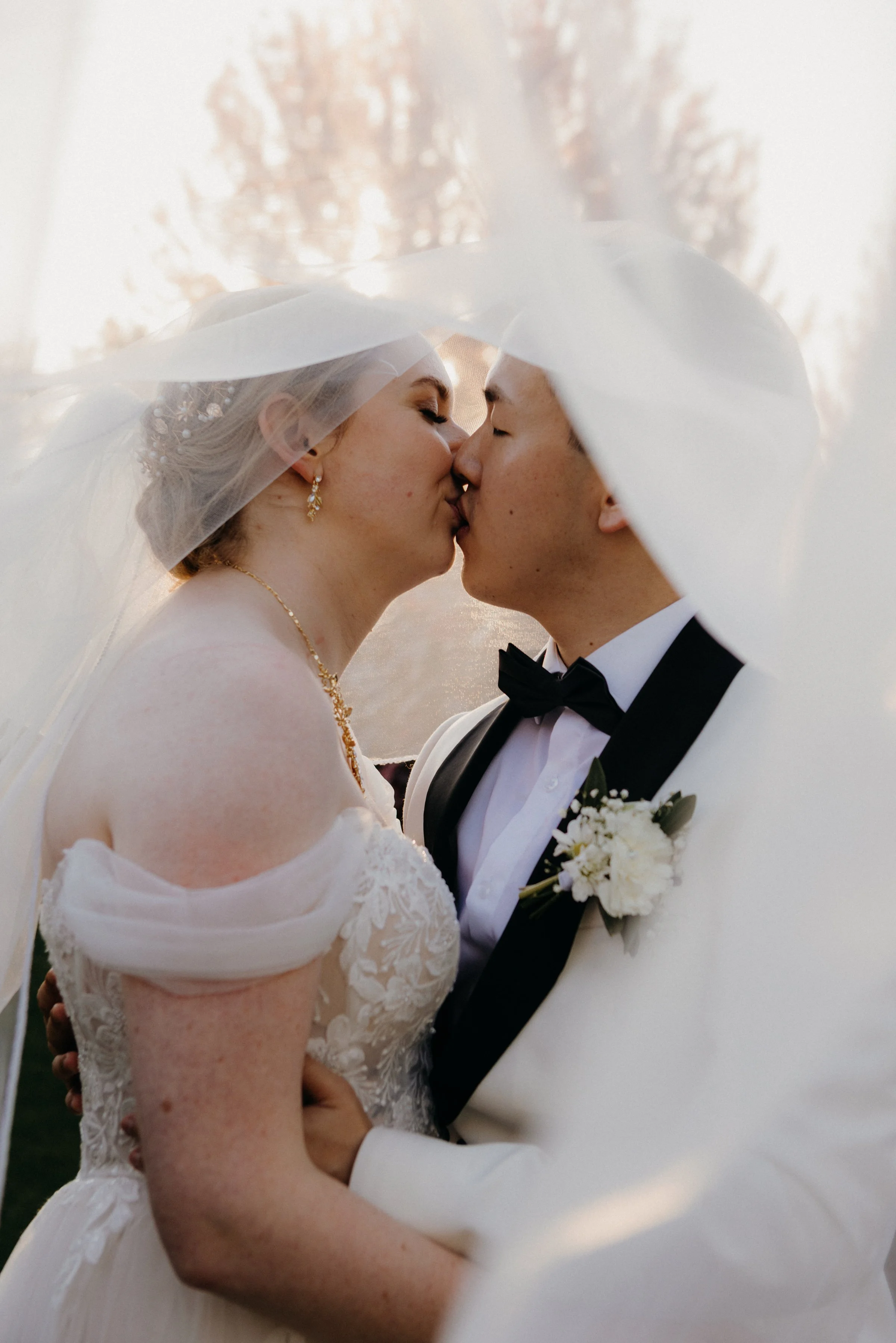 A bride and groom kiss under a veil during their wedding, outdoor setting with trees and sunlight in the background.