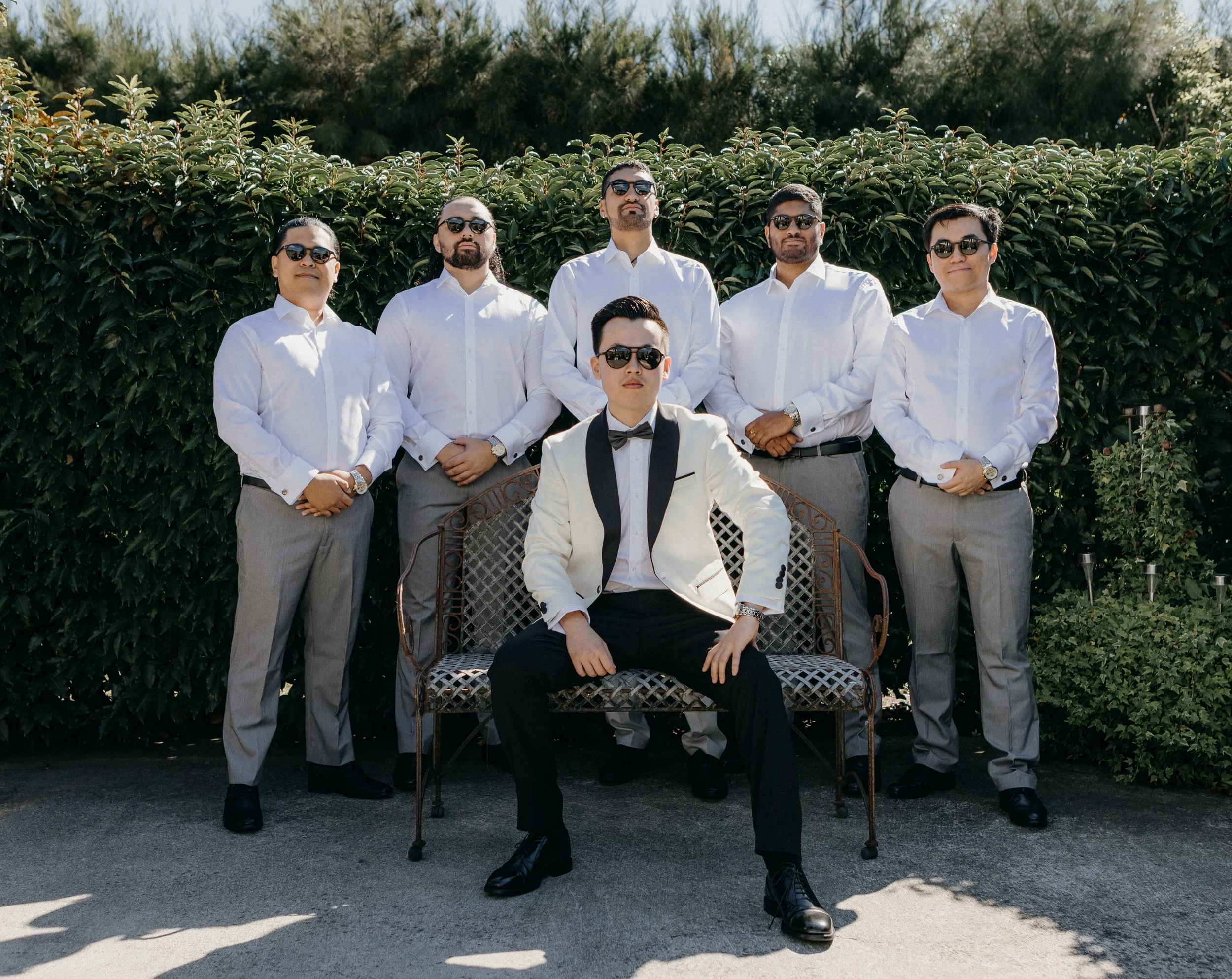A group of seven men dressed in formal attire posing outdoors in front of green bushes. The man in front is seated on a chair, wearing a white tuxedo jacket with black lapel, black bow tie, and black pants, with sunglasses. The other six men stand be