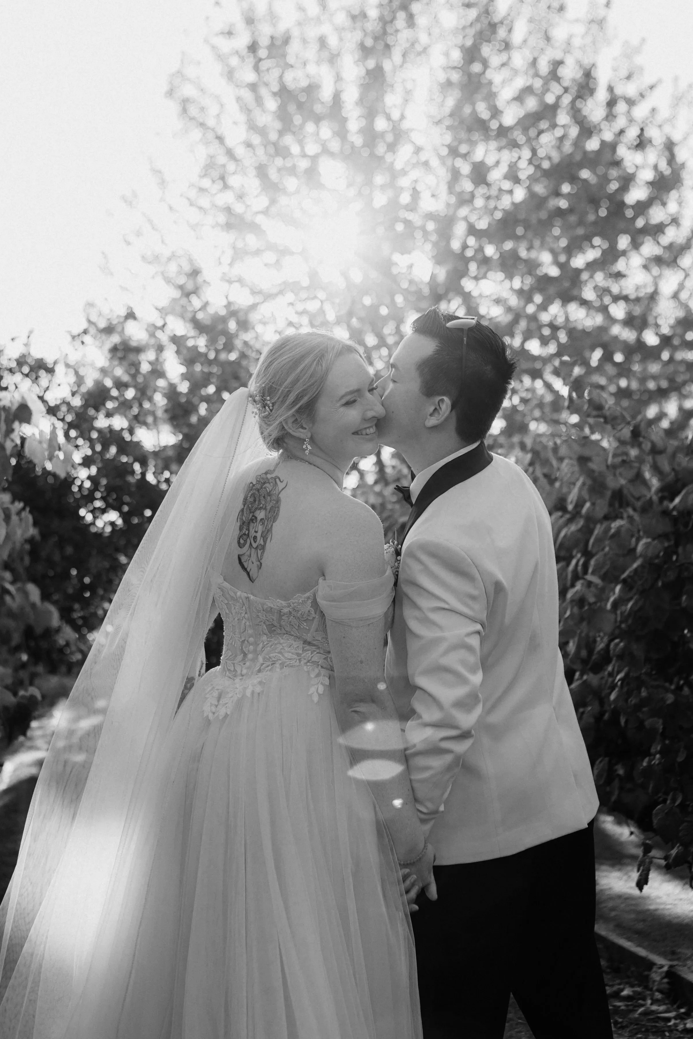 A bride and groom kiss outdoors at sunset, holding hands. The bride has a tattoo on her shoulder, wearing a wedding dress and veil. The groom wears a tuxedo. Sunlight filters through trees in the background.